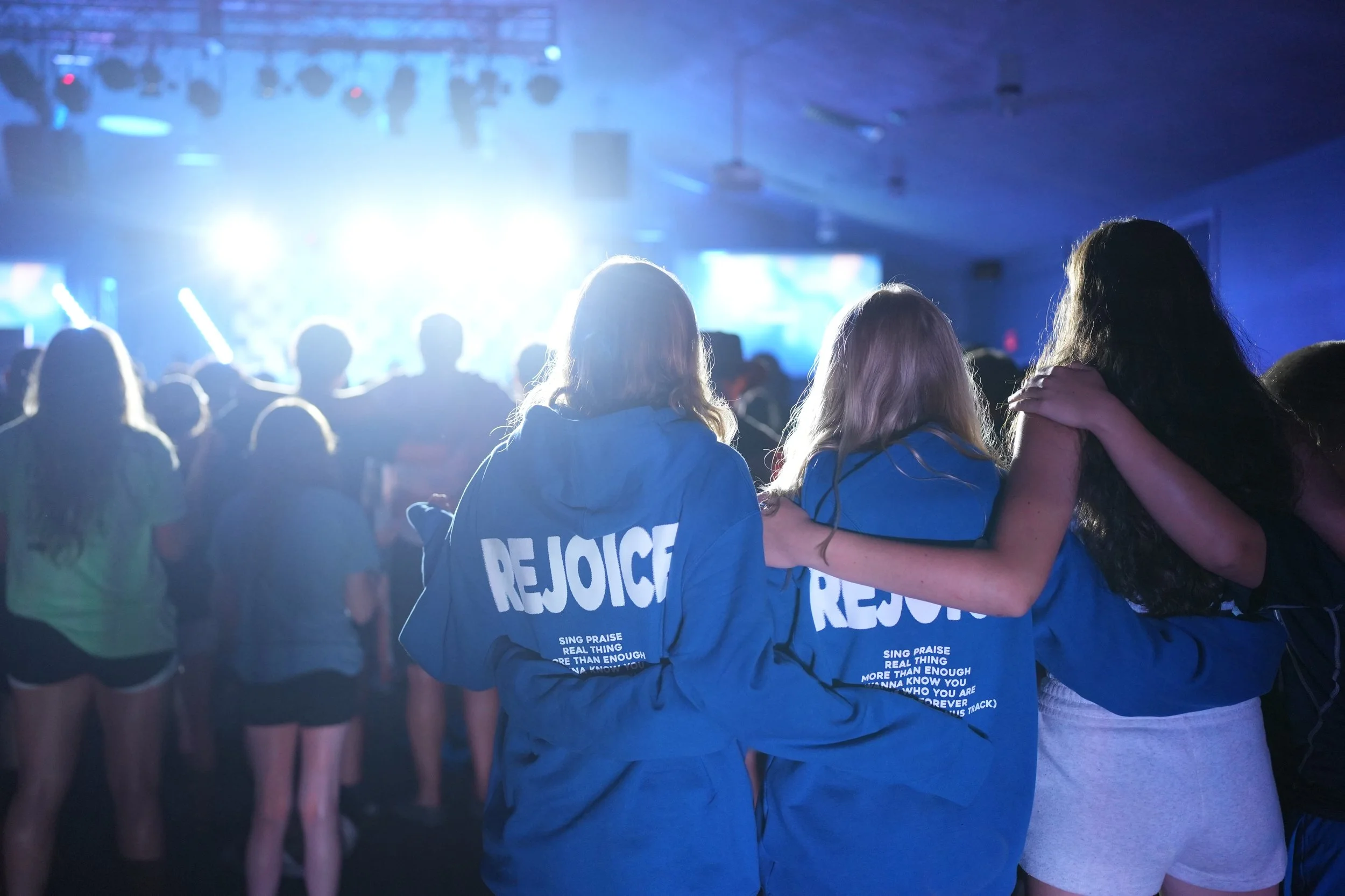 People at a concert with two women in blue jackets hugging in the foreground, stage lights in the background.