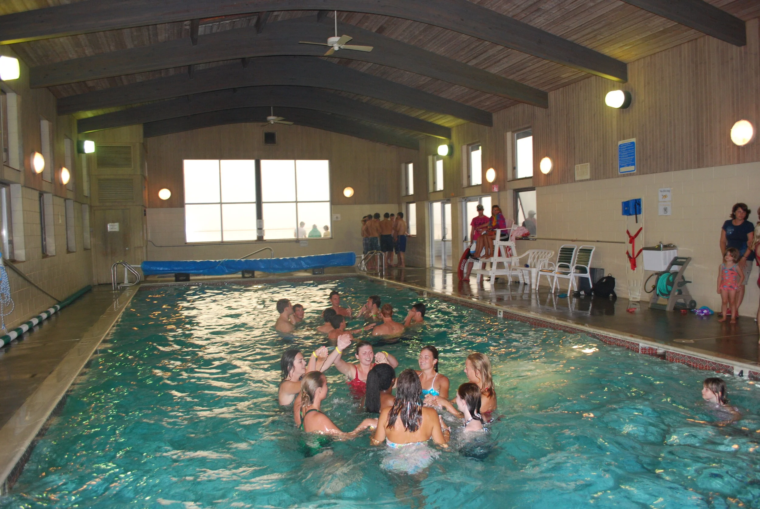 Indoor swimming pool with people swimming and socializing, lifeguards seated on chairs, and a group of children near the poolside, inside a wooden ceiling facility.