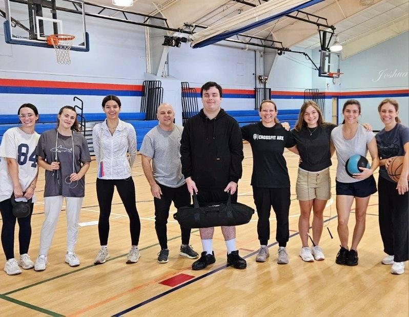 Group of nine people standing in a gymnasium with basketball hoops, some holding sports equipment, smiling for a photo.