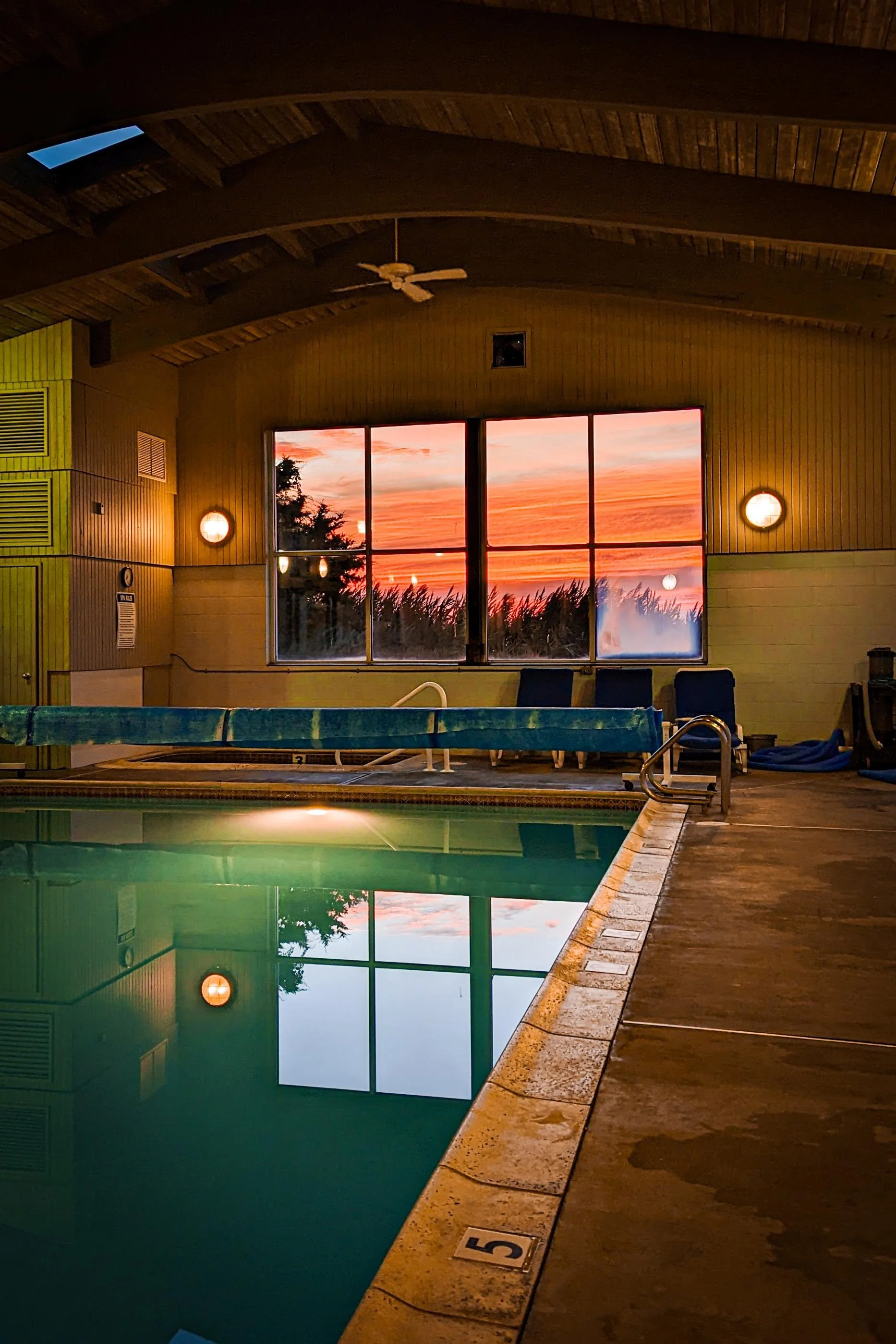 Indoor swimming pool with a sunset visible through large windows, illuminated by wall-mounted lights, with a ceiling fan and pool safety equipment.