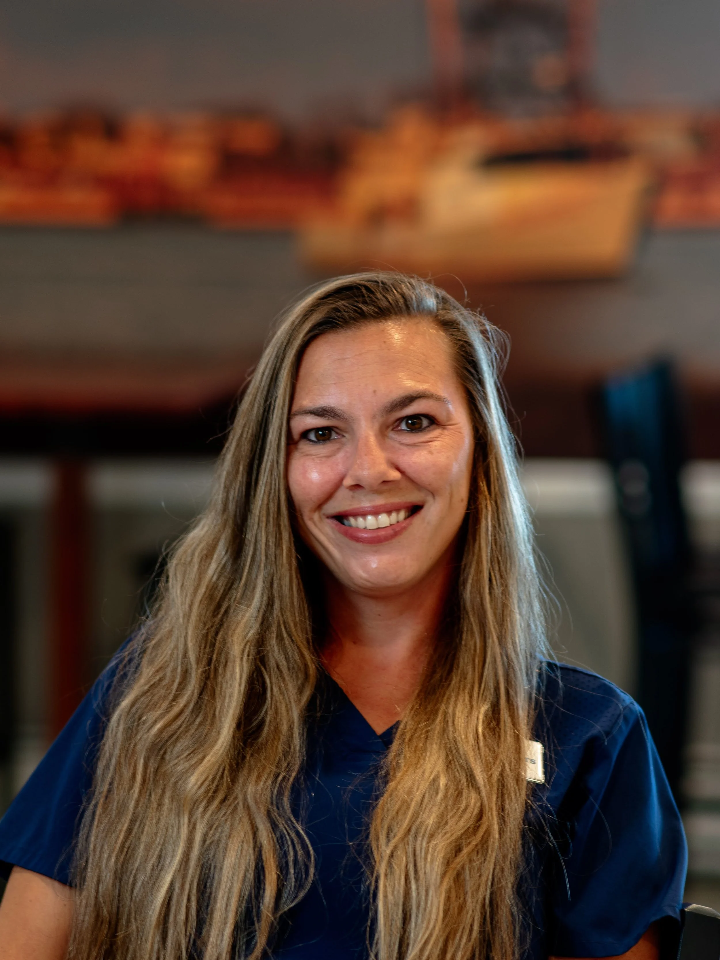 Close-up of a smiling woman with long brown hair tied in a bun, wearing a blue hoodie with 'Harvey Bible Camp' embroidered on it, indoors with a blurred background.