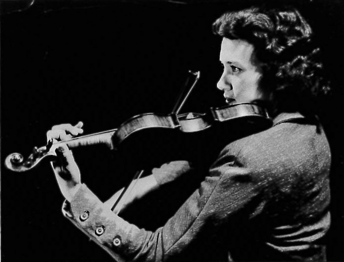 A black and white photo of a woman with curly hair playing a violin, facing to the right.