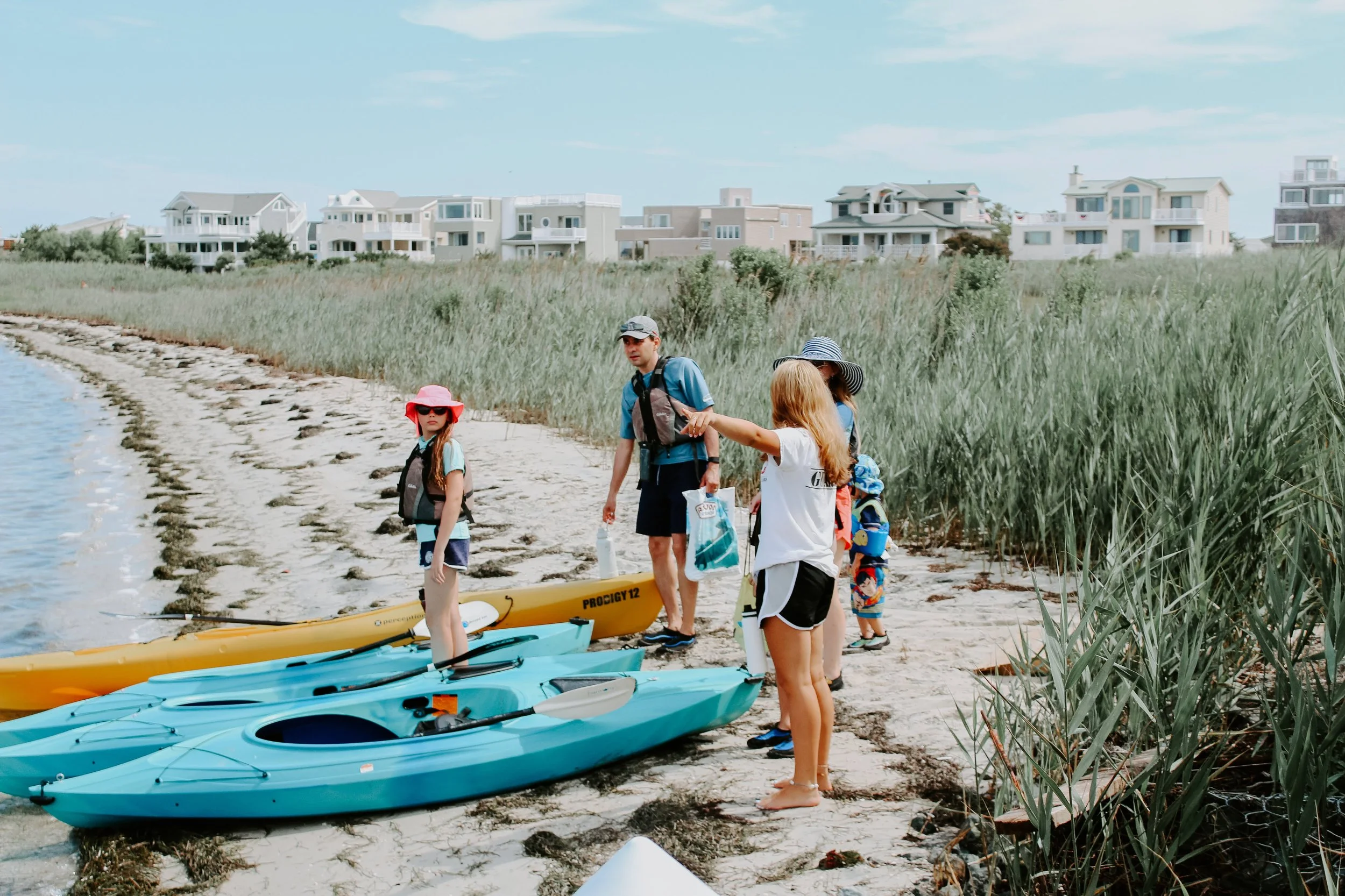Group of people preparing to kayak on a sandy beach near tall grass and beachfront houses in the background.
