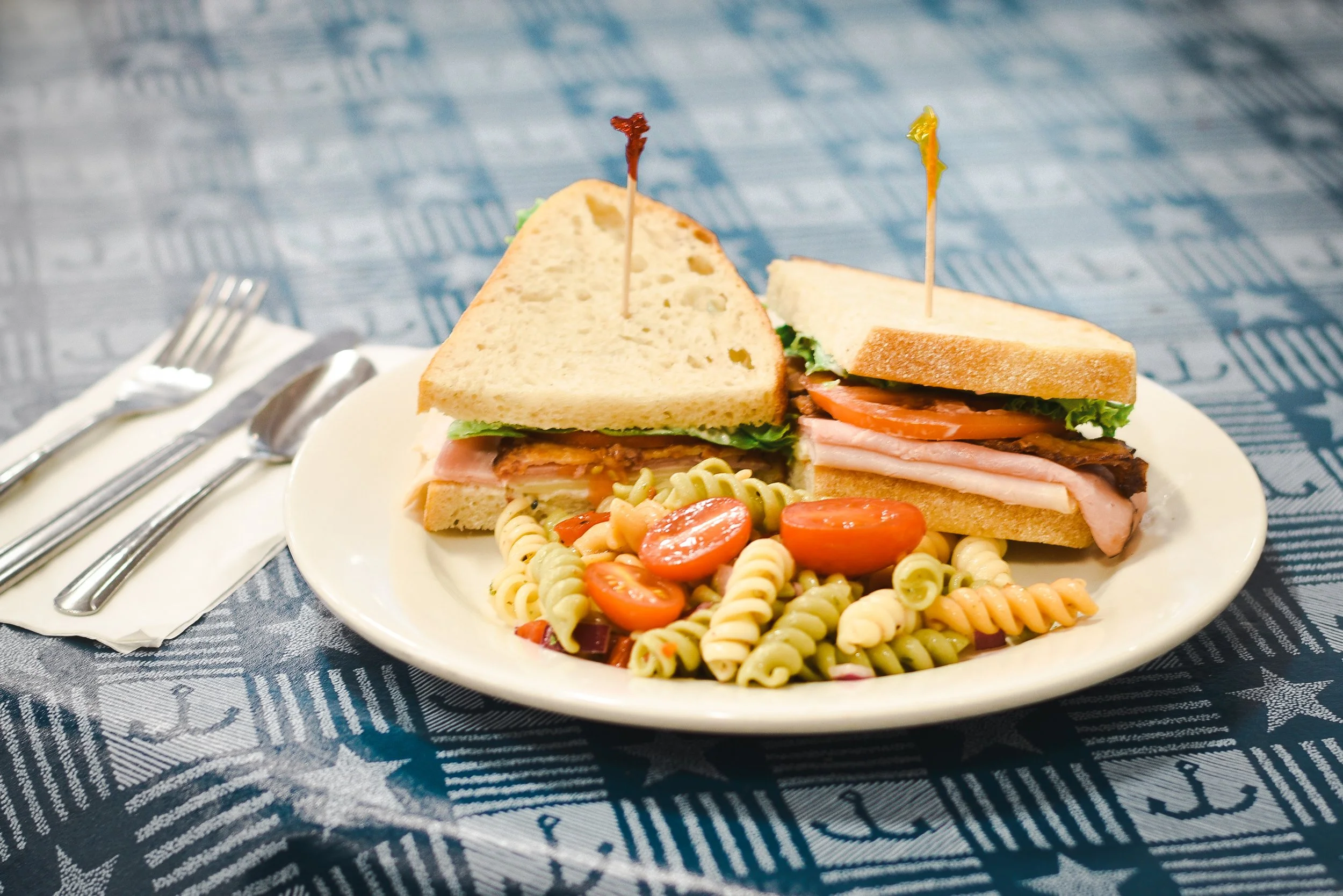 A plate with two sandwiches, spiral pasta salad with cherry tomatoes, and a fork and knife on a napkin, on a table with a blue cloth featuring white star and stripe patterns.