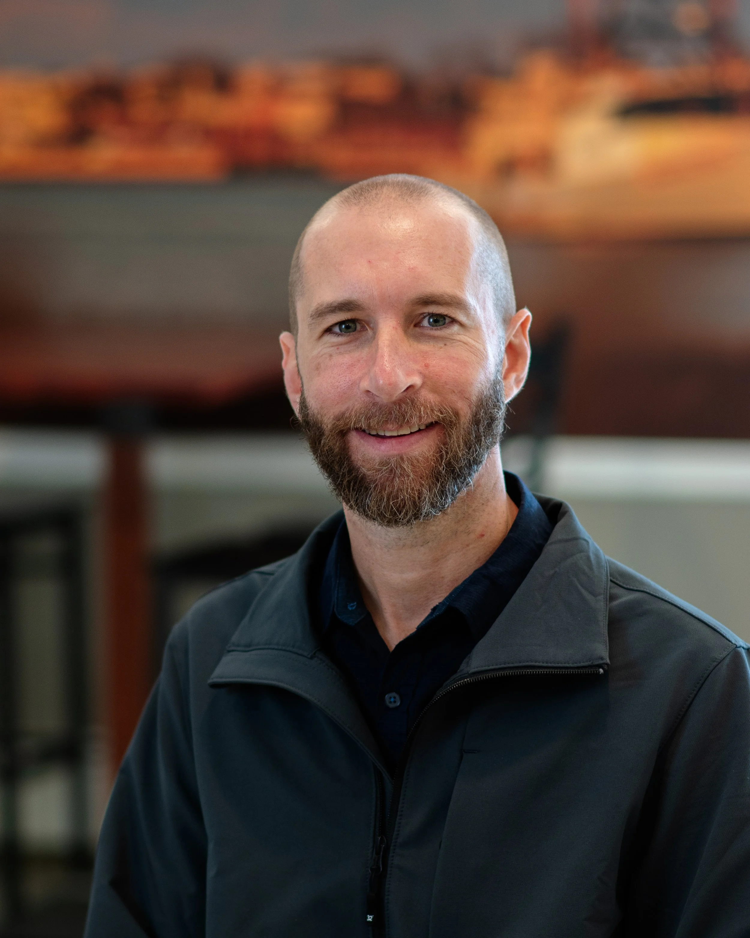 A man with a shaved head and beard, smiling, wearing a black jacket and navy shirt, standing indoors in front of blurred artwork or photograph.