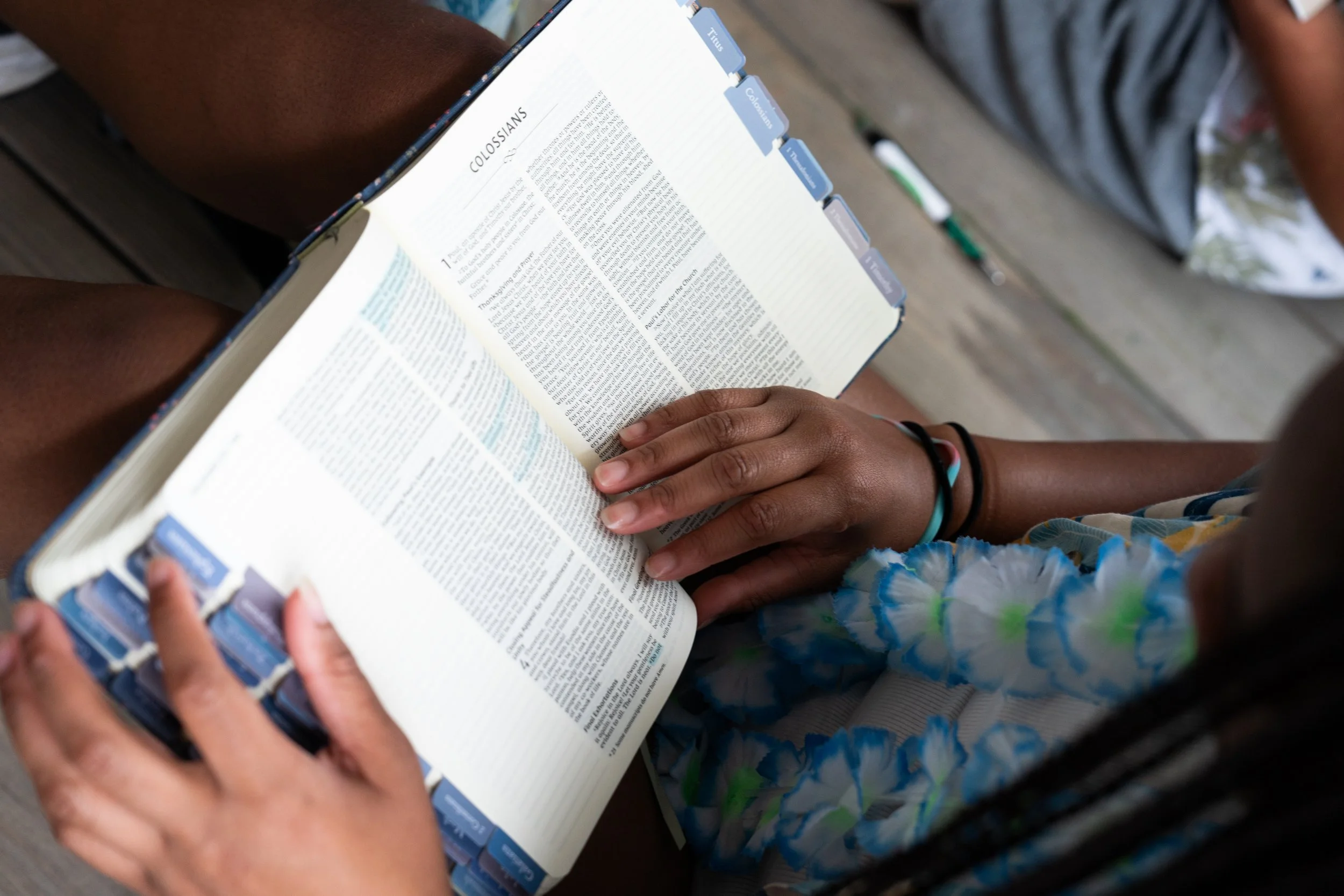 A person holding a large open Bible with tabs marking different sections, including titles like "Titus," "Cohesion," and "1 Timothy." The individual is sitting outdoors, dressed in a striped shirt and a blue, green, and yellow tie-dye skirt, with bracelets on their wrist, and is resting their hand on the Bible.