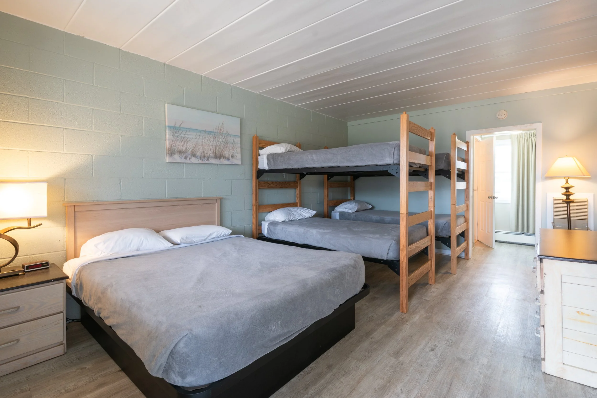 A cozy bedroom with a double bed and a bunk bed. The room has light-colored walls, a wooden nightstand with a lamp, and a piece of wall art depicting beach grass. There is a window with curtains letting in natural light, and the flooring appears to be wood or laminate.