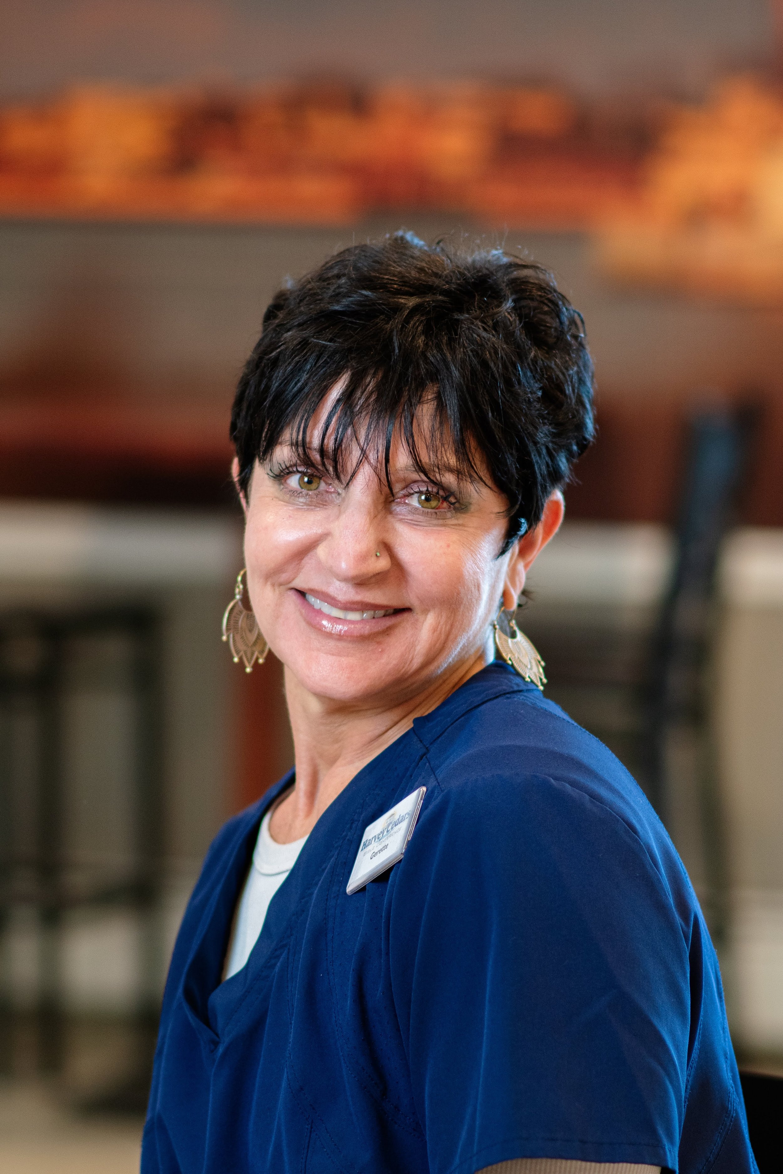 A woman with short dark hair, wearing blue medical scrubs, smiling at the camera in a professional setting.