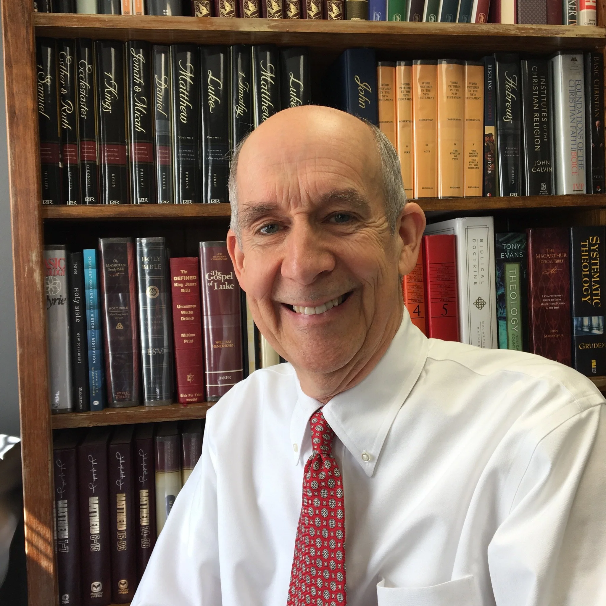 Part of a smiling man's face, showing forehead, right eye, and ear, with a background of bookshelves filled with religious and theological books.