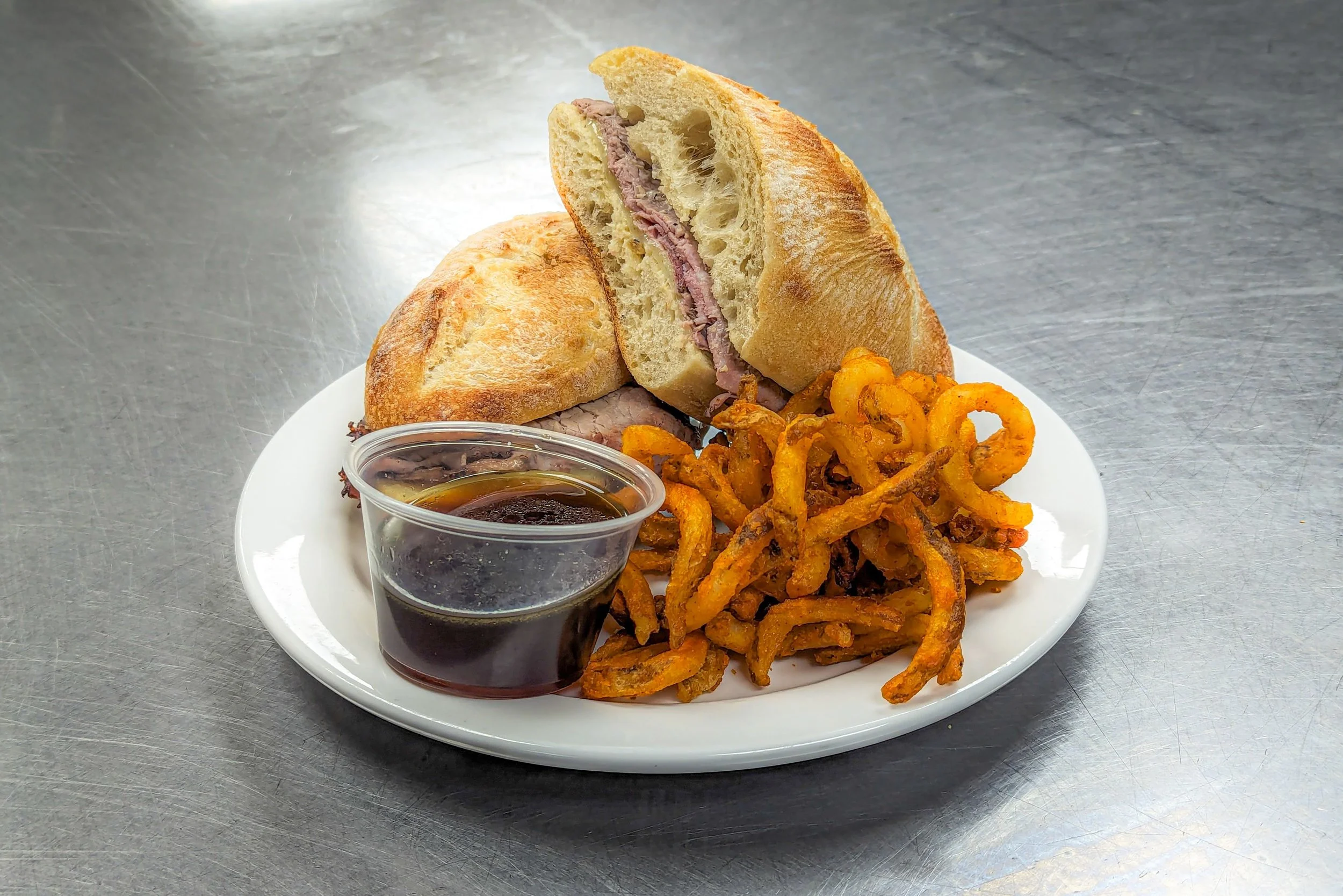 Plate with sandwich, fried onions, and small container of dark dipping sauce on a metallic surface.