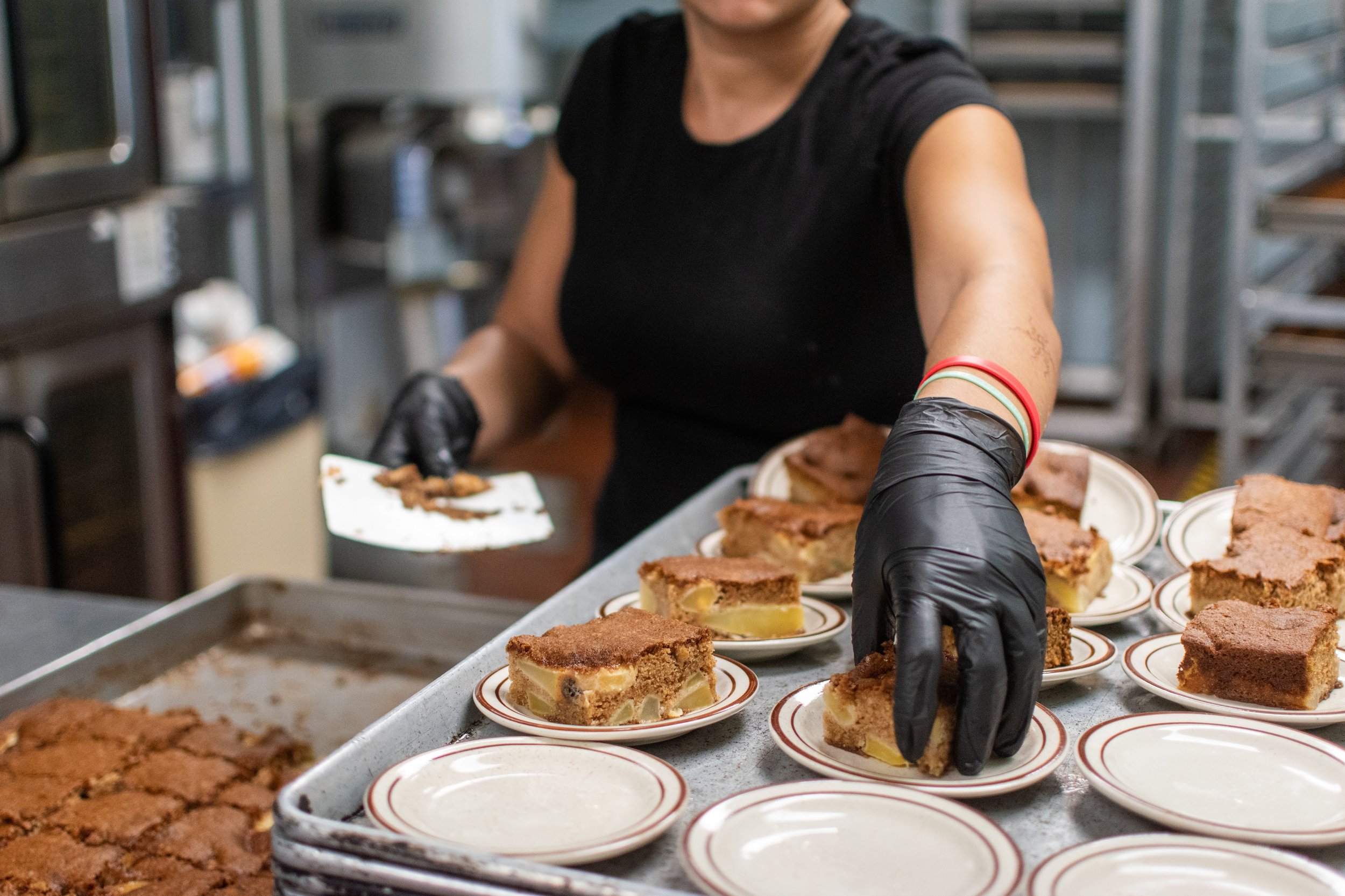 A person wearing black gloves and a black shirt arranging slices of pie on small plates in a kitchen or bakery setting.