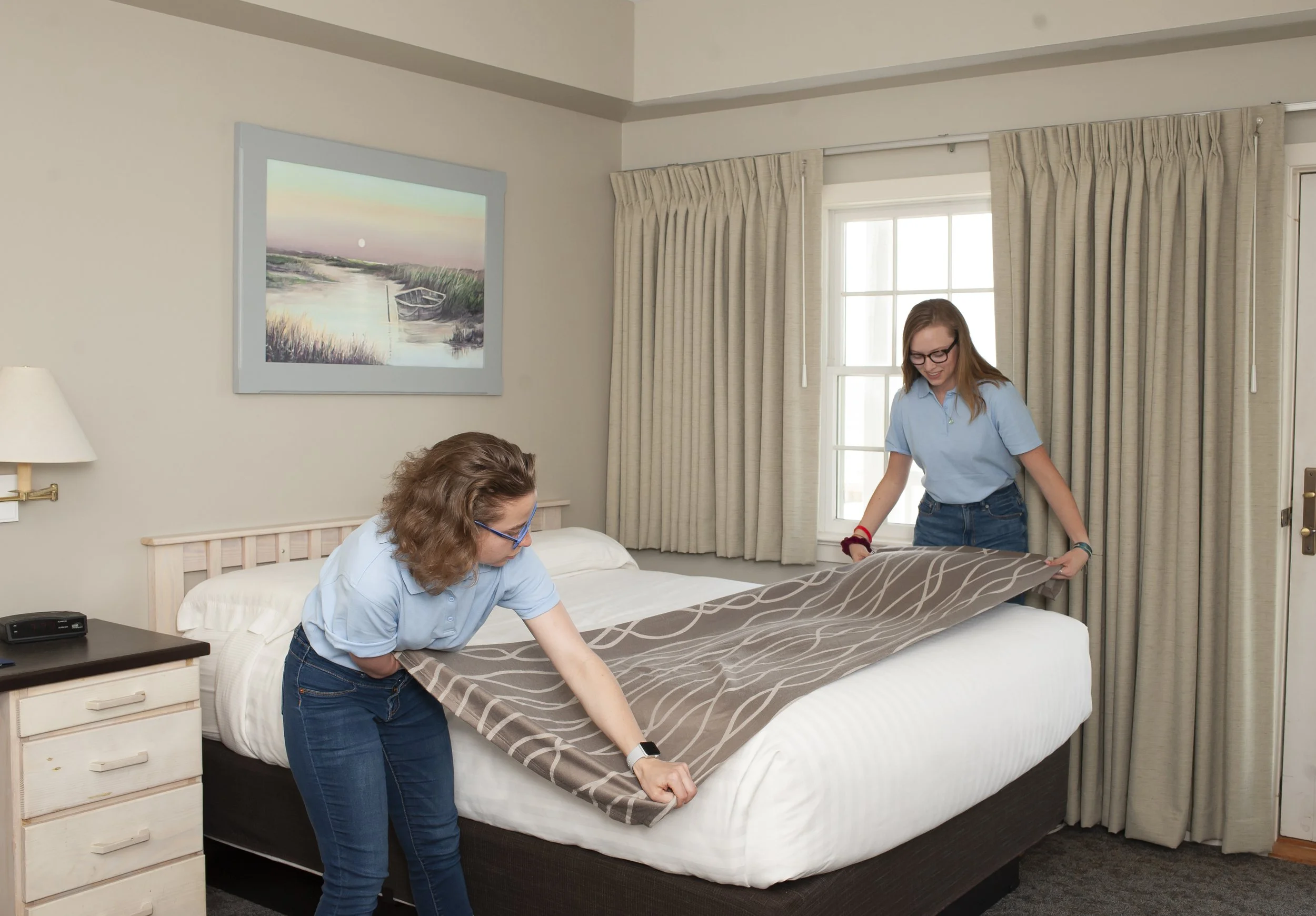 Two women are making a bed together in a hotel room, one is folding the blanket and the other is smoothing it out. They are both wearing light blue shirts and jeans.
