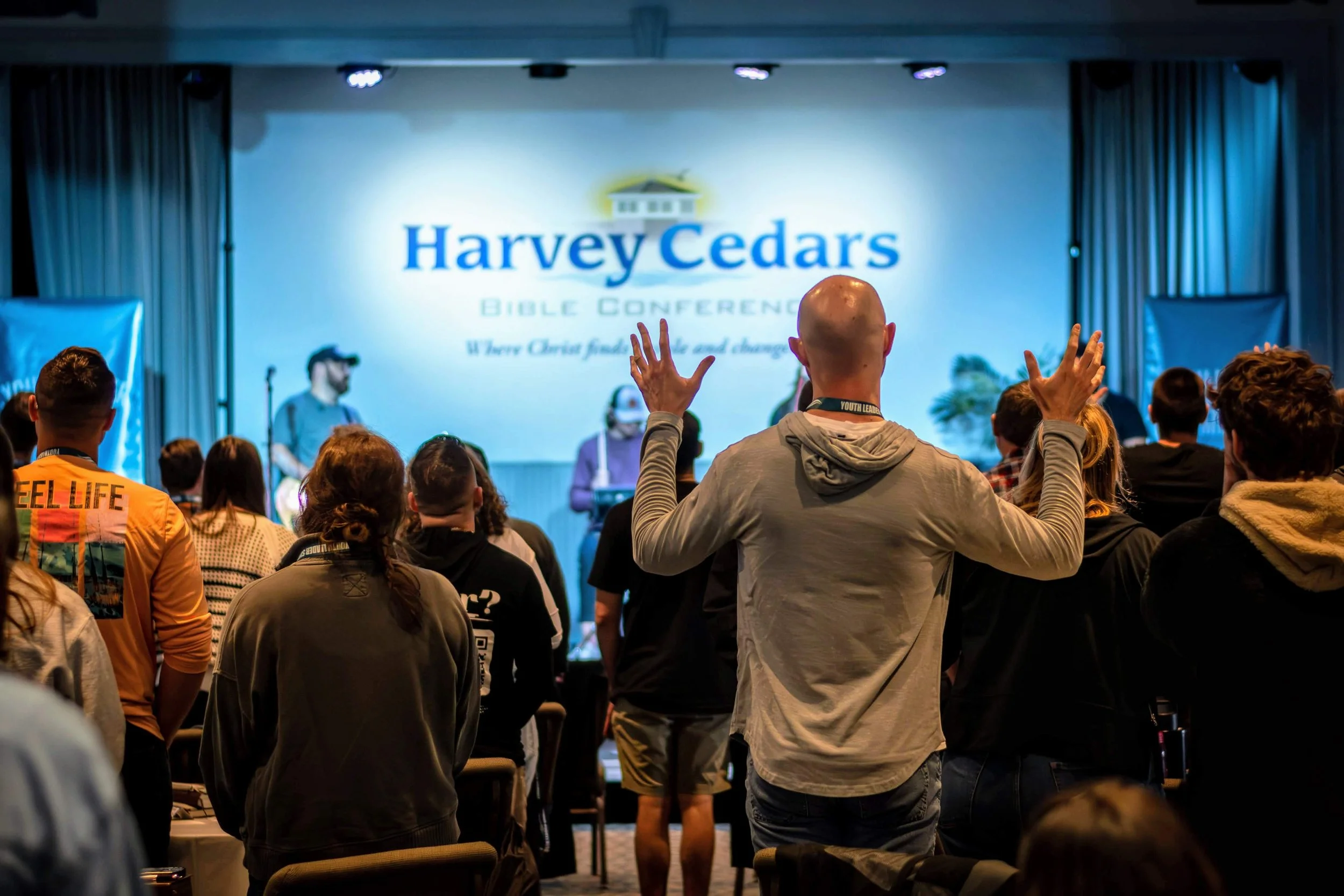 Audience at Harvey Cedars Bible Conference raising their hands during a presentation in a large room with a stage and a screen displaying the conference logo.
