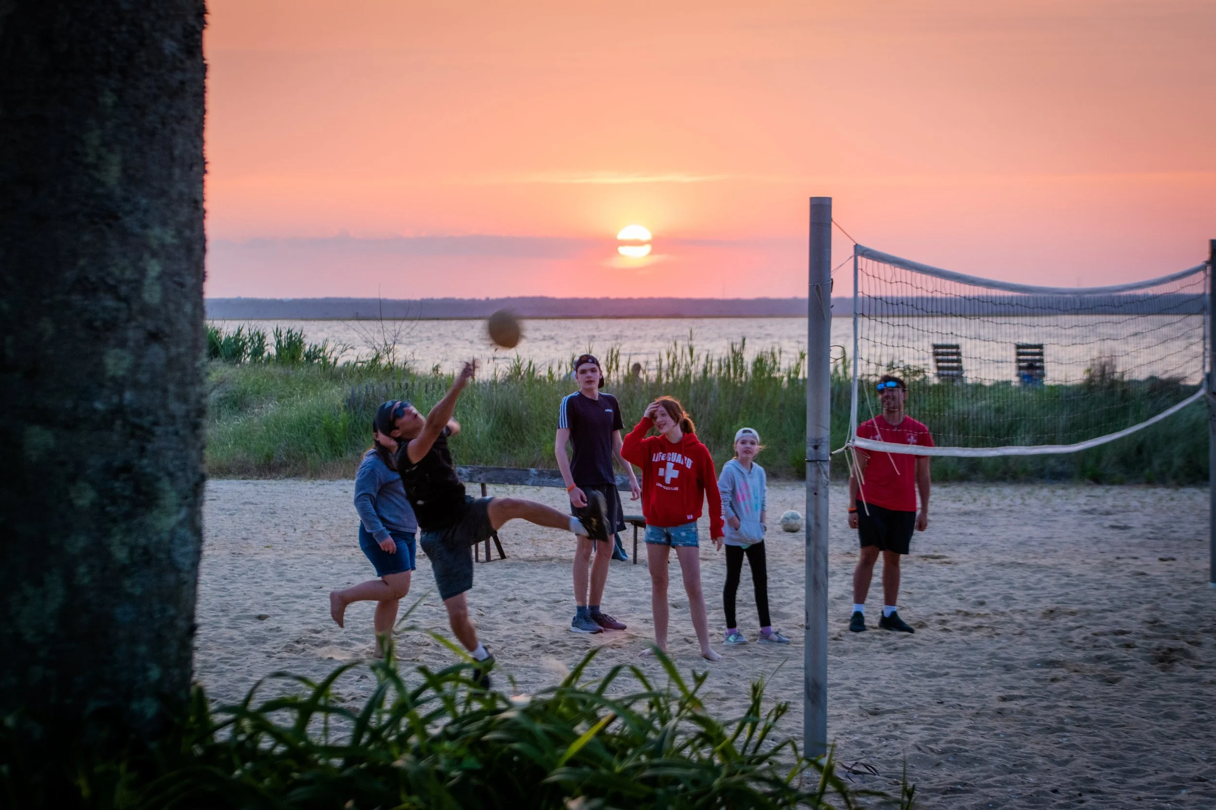 A group of people playing volleyball on a sandy beach at sunset, with a body of water and a sunset in the background.