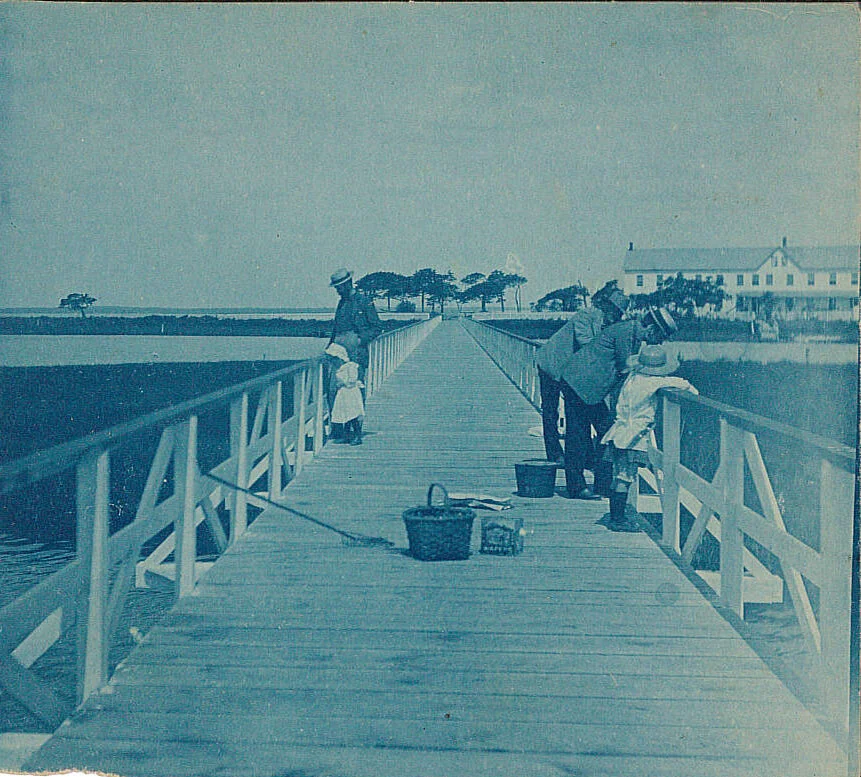 Black and white photo of people fishing off a wooden pier with a house and trees in the background.