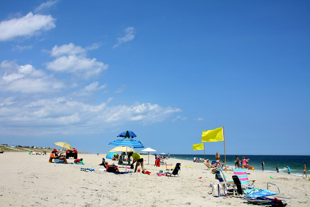 Beach scene with umbrellas, chairs, and people enjoying the sun and ocean under a partly cloudy sky.