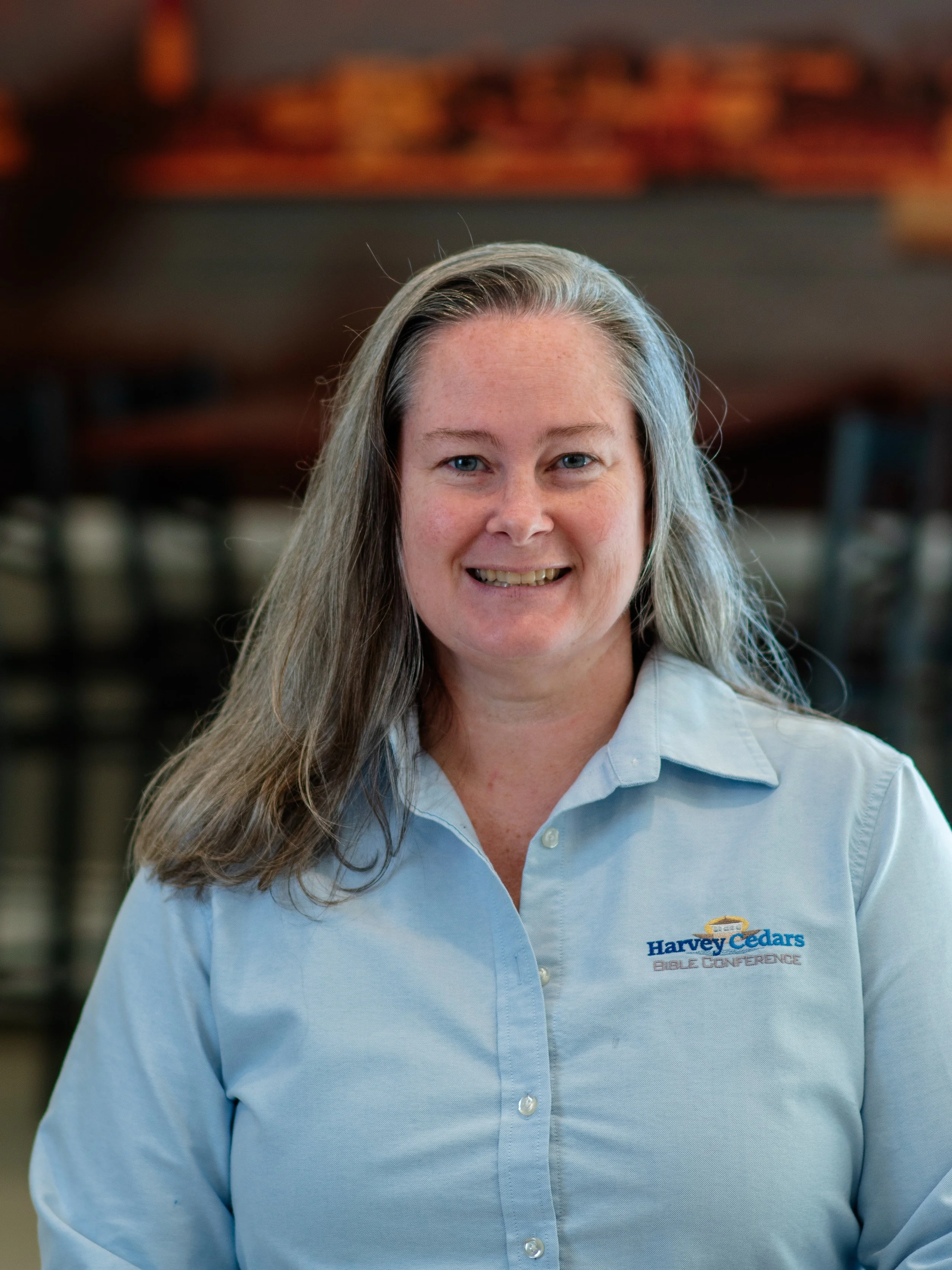 A smiling woman with long, light brown hair, wearing a light blue button-up shirt with a logo that says Harvey Cedars Bible Conference, standing indoors with blurred background of bookshelves.
