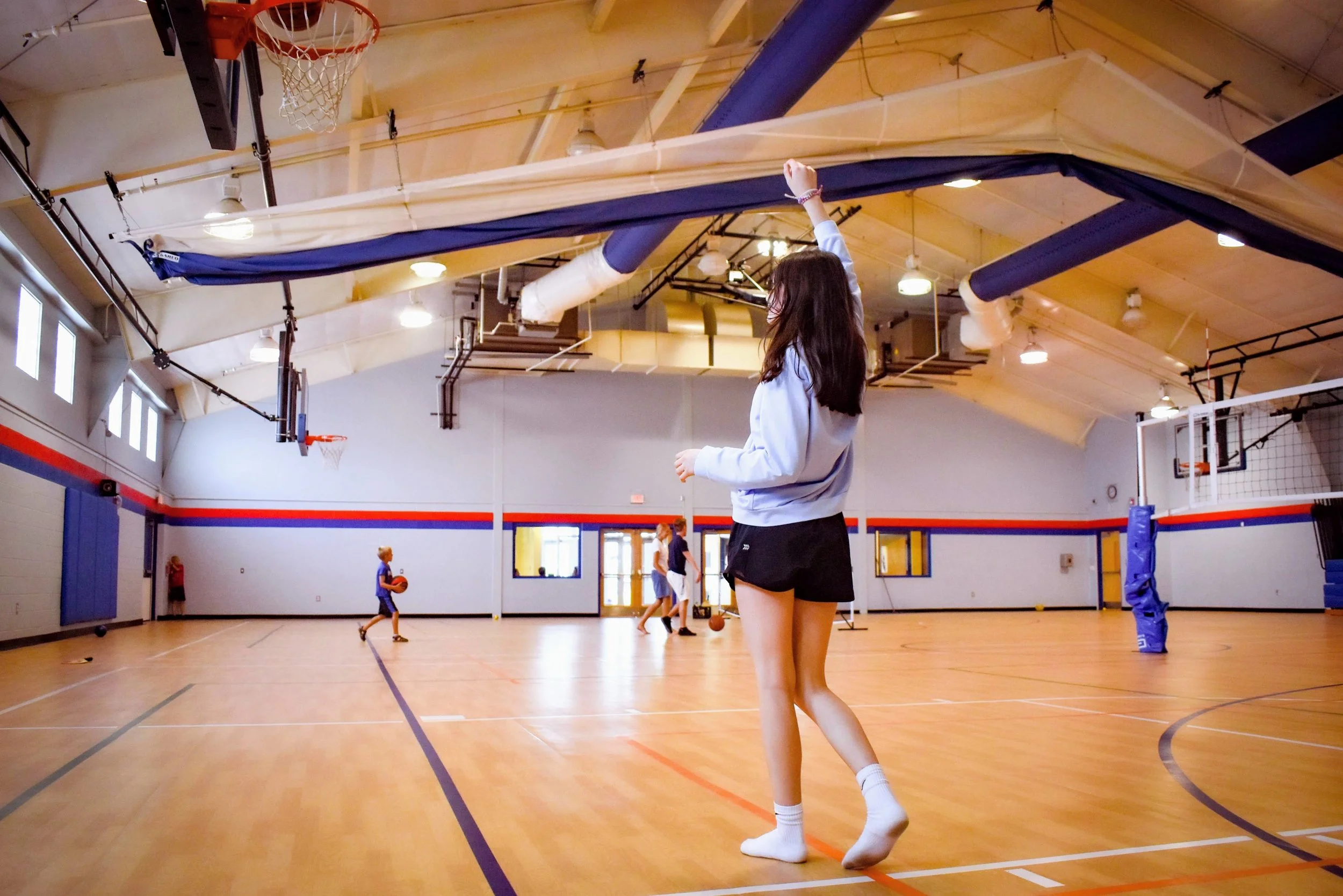 Young girl playing basketball in an indoor gymnasium, with other kids in the background also engaging in basketball activities.