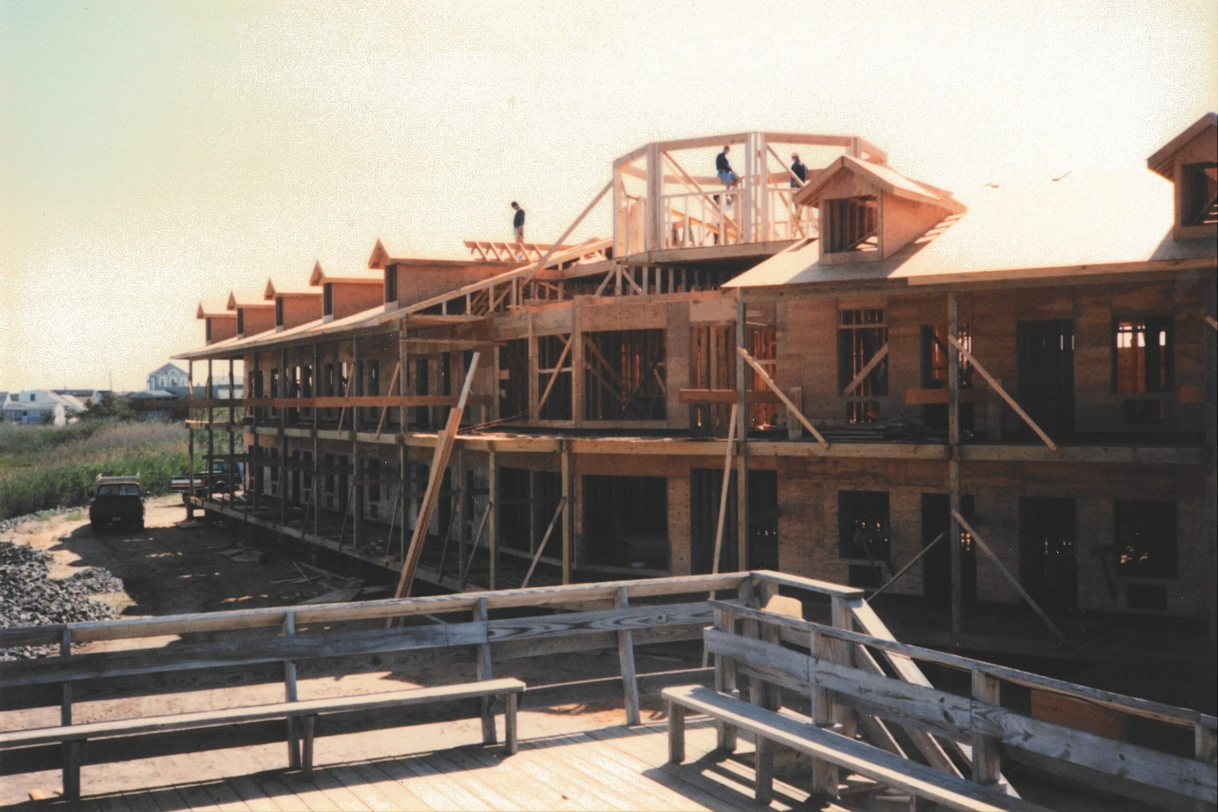 Construction site of a multi-story wooden building, with workers on the roof framework and scaffolding around the structure, during daytime.