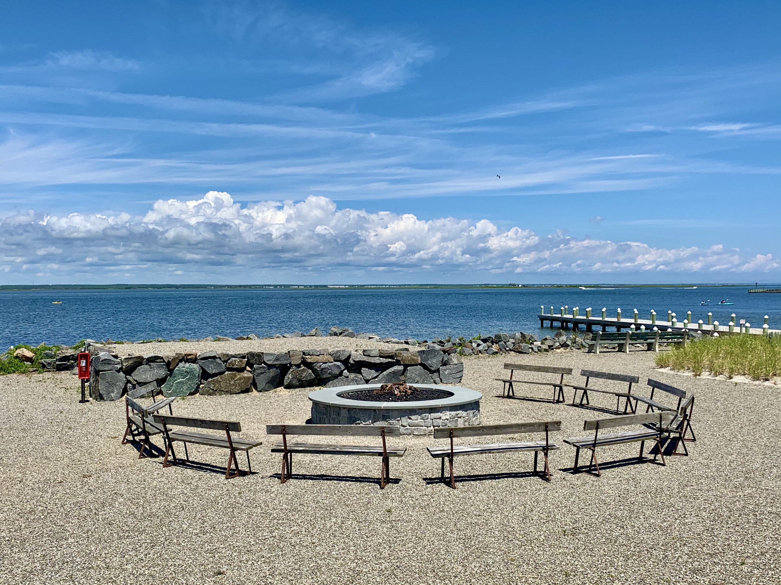 Beachside with a circular fire pit surrounded by benches facing the water, a rocky barrier, and a pier extending into the ocean under a partly cloudy sky.