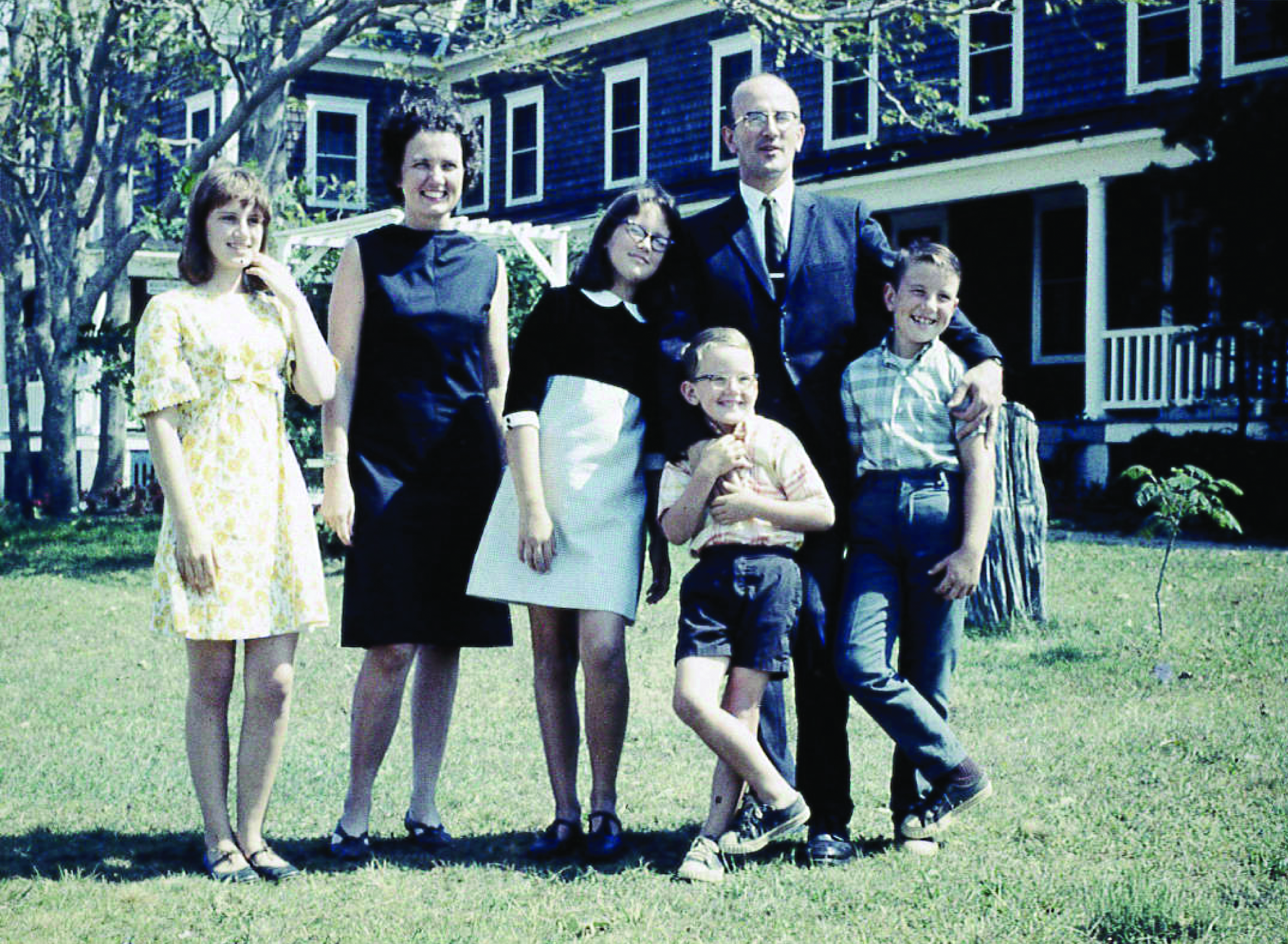 A group of seven people standing on a lawn in front of a house with blue siding and white trim, smiling, including children and adults.
