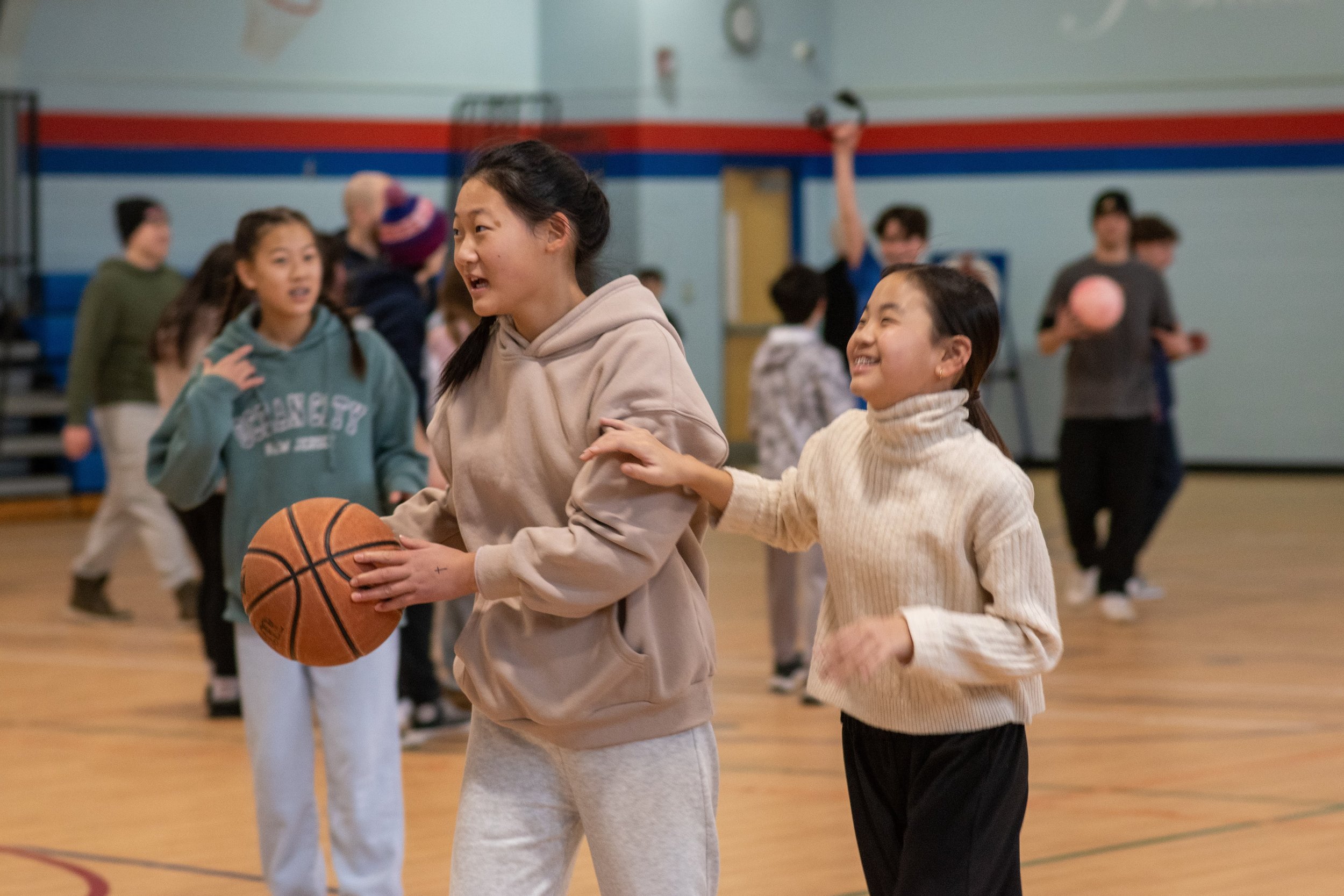 Two young girls are playing basketball in a school gymnasium, with other kids in the background.