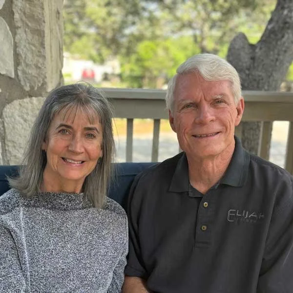 A smiling senior woman and man sitting outdoors on a porch with a tree and greenery in the background.