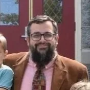 A smiling man with glasses and a beard wearing a brown blazer, pink shirt, and patterned tie. Part of a child's arm is visible on his shoulder, and they are outdoors with a red door and stone wall in the background.