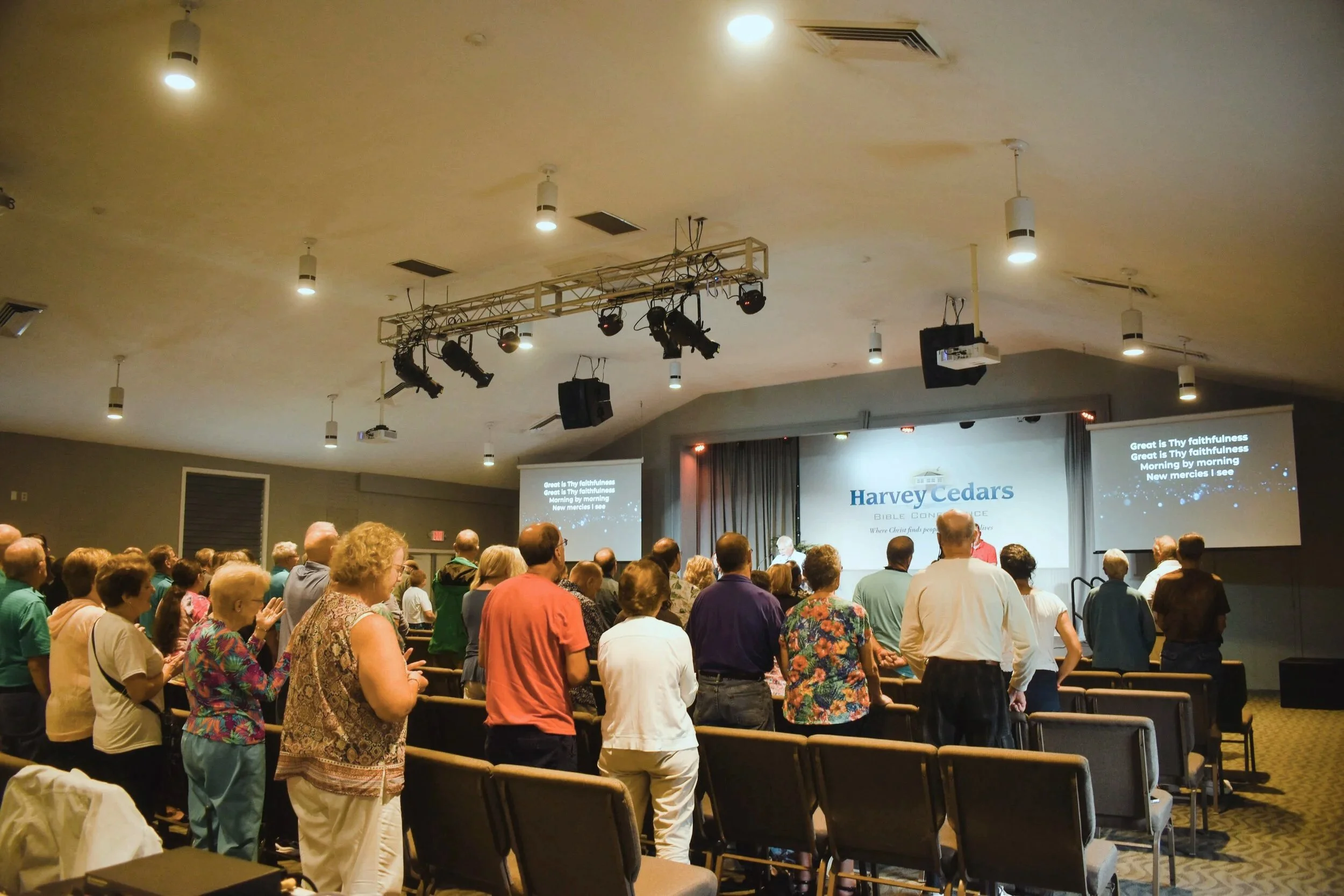 A group of people standing in a church or conference hall, facing forward and participating in a religious service or event. The room has a ceiling with hanging lights, speakers, and video screens displaying lyrics and the name 'Harvey Cedars'.