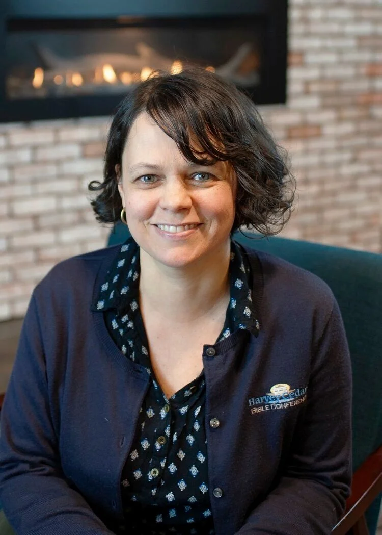 A woman smiling and sitting indoors with a brick fireplace in the background, wearing a dark blue jacket with a logo that reads Harvey Cedar Bible Conference.