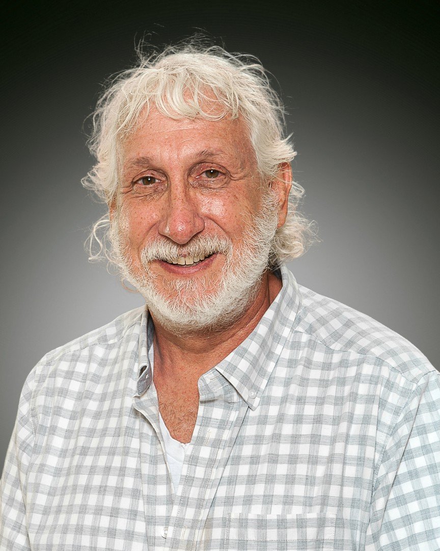 Portrait of an older man with white curly hair, a beard, and a mustache, smiling, wearing a light-colored checked button-up shirt, against a dark gray background.