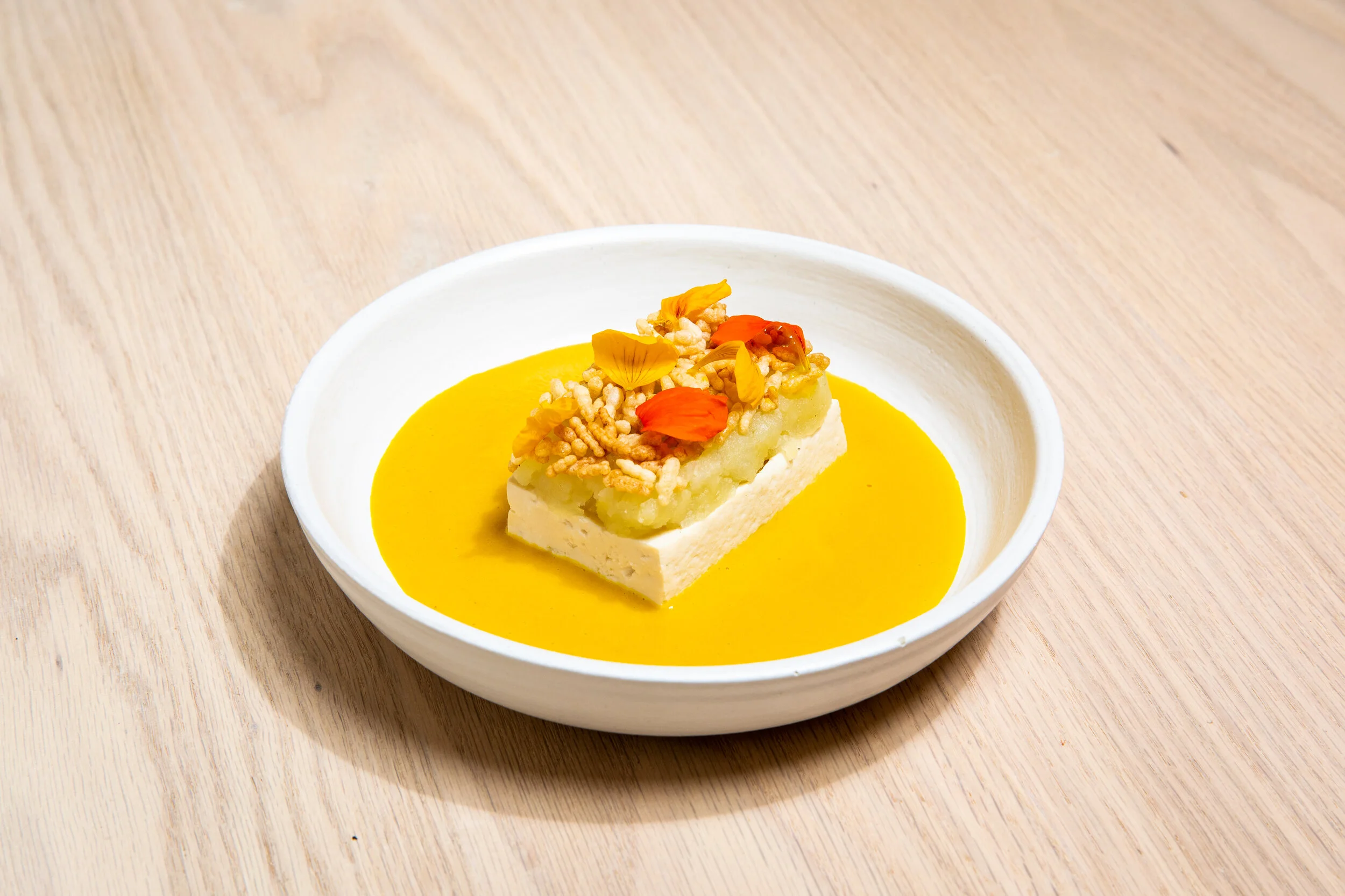 A dish with a rectangular piece of white cheese or tofu, topped with yellow and orange flower petals and crunchy rice cereal, served on a bed of bright yellow sauce in a white bowl on a light wooden surface.