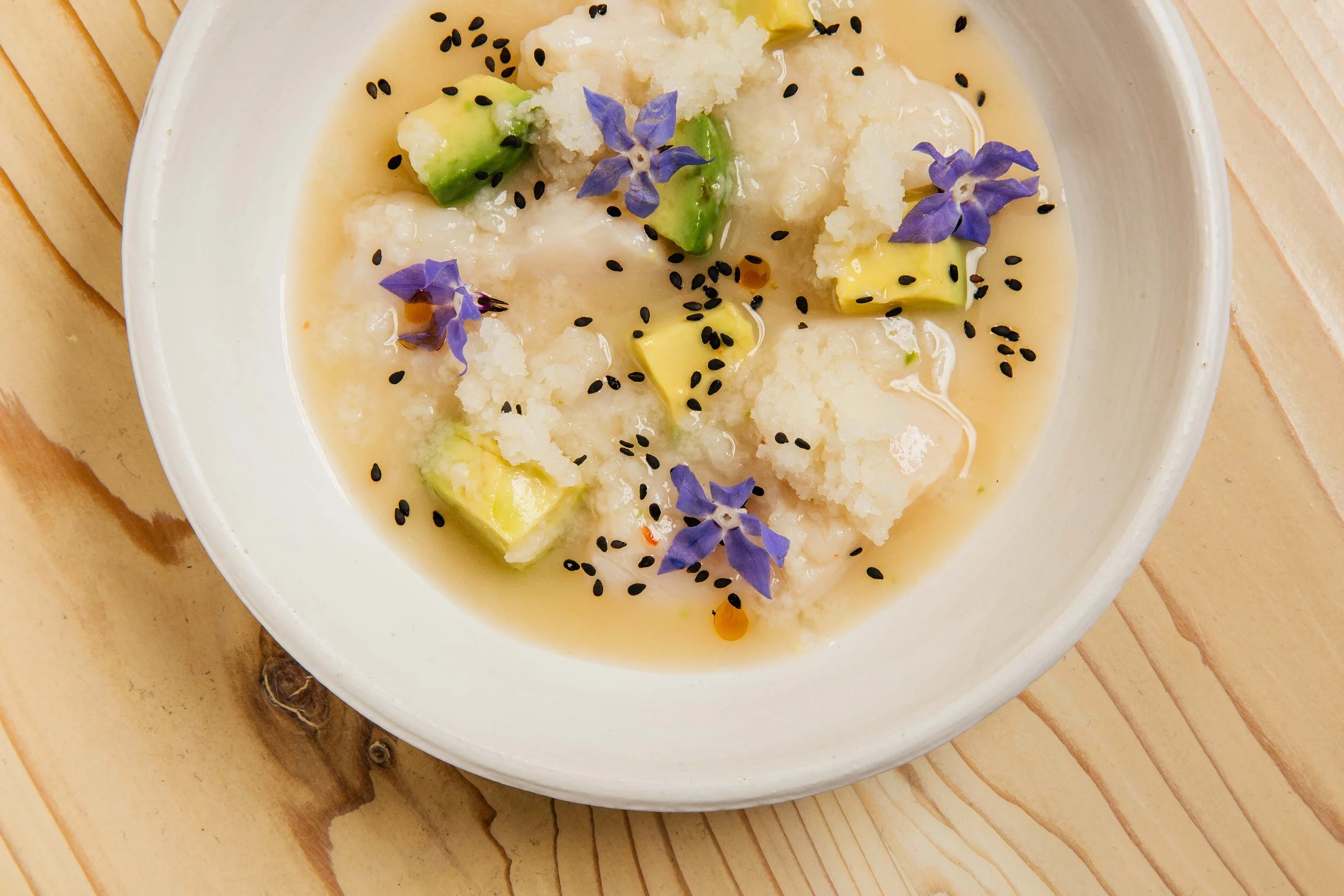 Close-up of a dish with cubed avocado, grated cheese, black sesame seeds, and purple edible flowers in a creamy sauce served in a white bowl on a wooden surface.