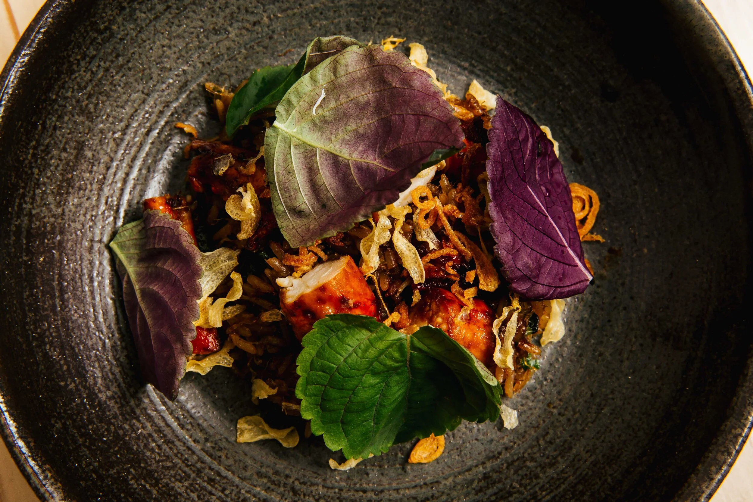 Close-up of a gourmet dish with cooked meat, garnished with purple and green edible leaves, crispy fried onions, and herbs, served on a dark textured plate.