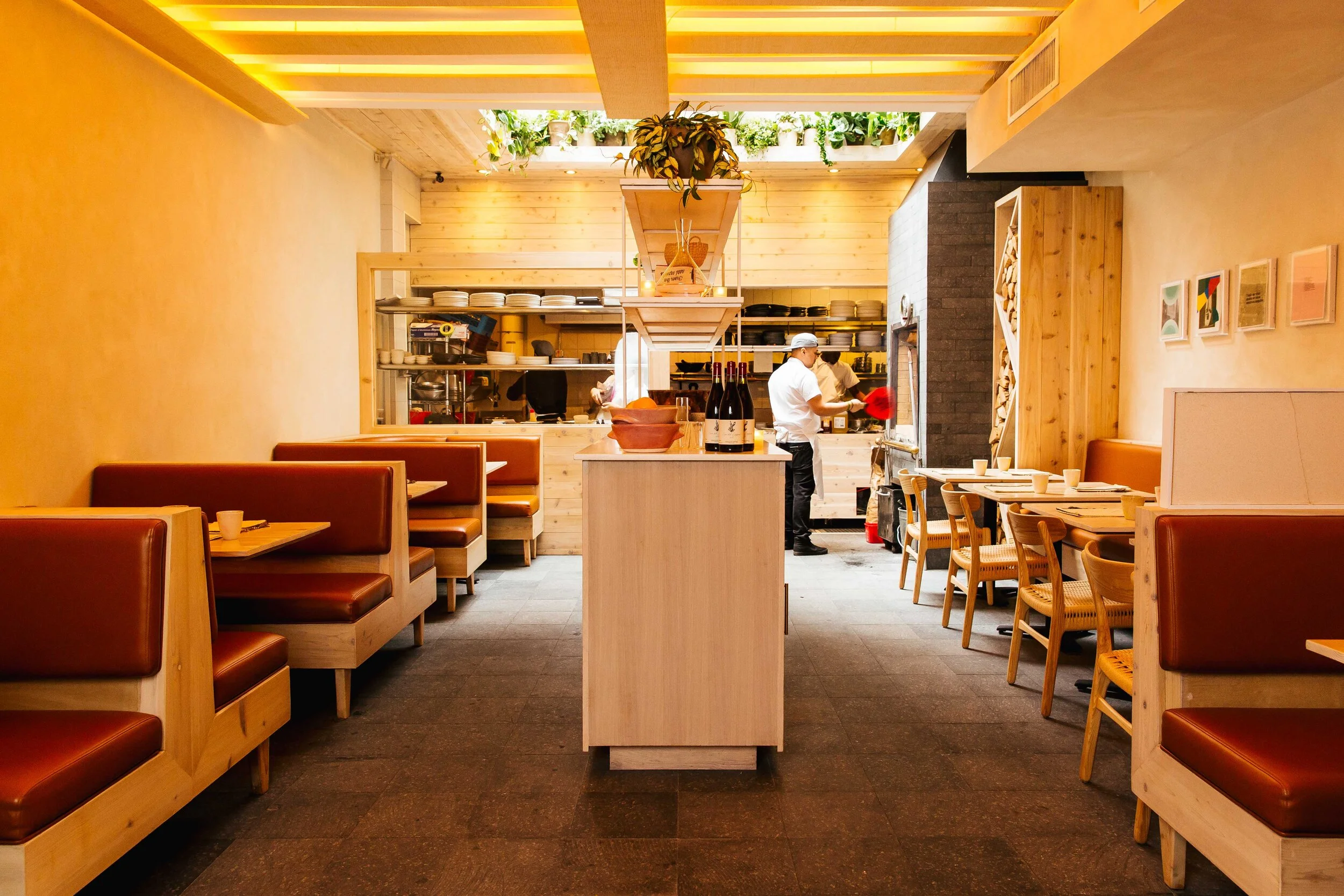 Interior of a modern restaurant with wooden decor, booths on the left and tables on the right, a kitchen open to the dining area in the background, and chefs preparing food.