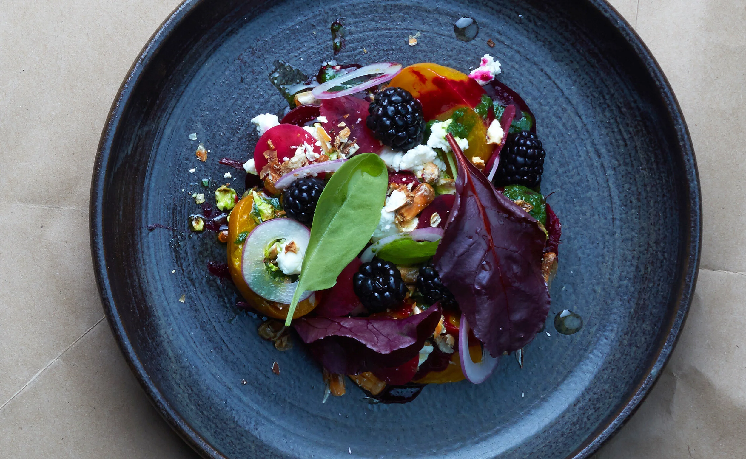 A colorful salad with mixed greens, blackberries, tomatoes, red onions, nuts, and cheese on a dark ceramic plate.