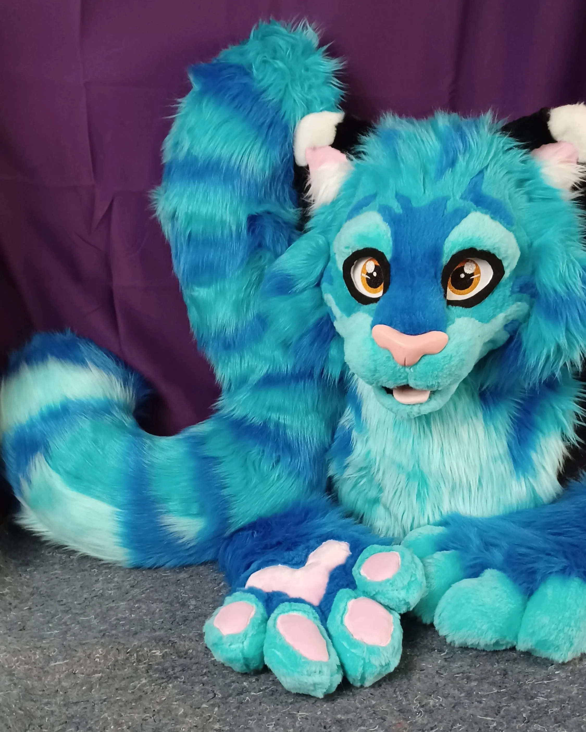 Colorful furry blue, green, and black unicorn mascot costume with large eyes, pink nose, and pink paw pads, sitting on a carpeted floor near a purple curtain.