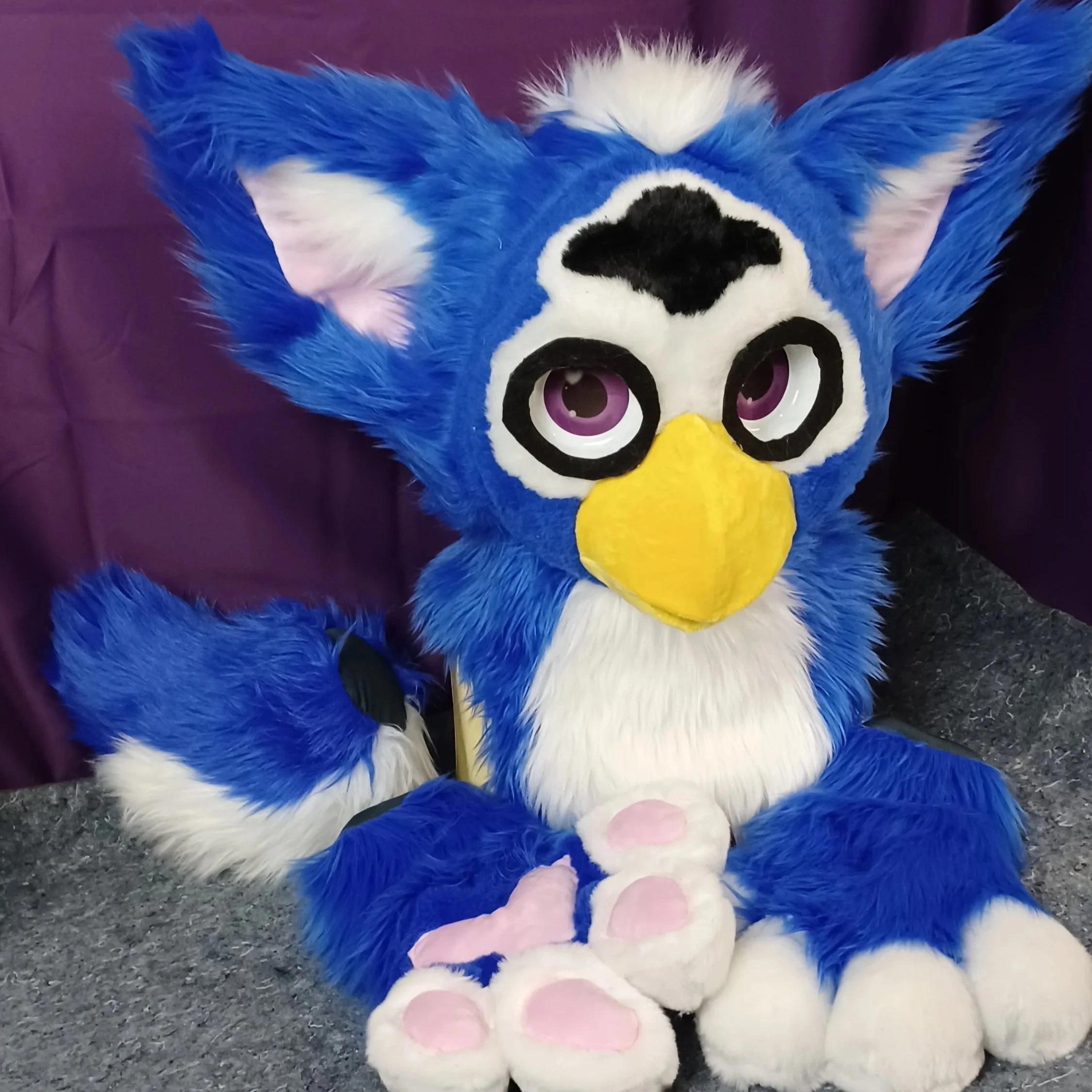 a furby costume with blue, white, black, fur, purple eyes, and a yellow beak, with a purple background.