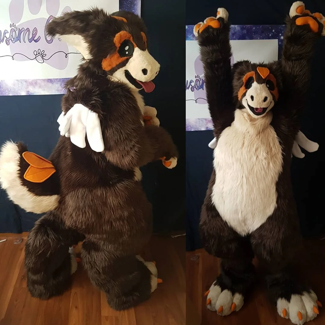 A person in a large, furry, angel dragon costume with a big white belly, orange and black facial markings, and small wings on the arms, standing inside a room with wooden flooring and a dark background.
