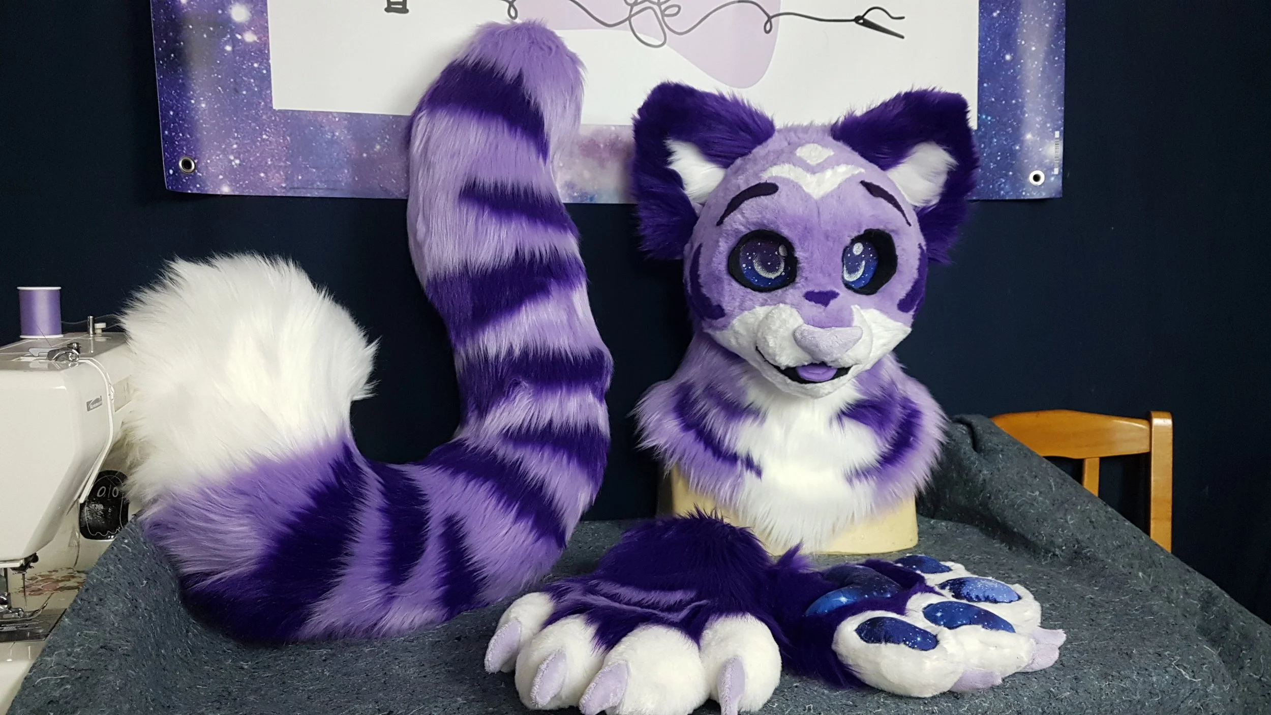 Purple and white furry tiger costume with big eyes and striped tail sitting on a table, with another similar tail visible in the foreground, against a starry galaxy background.