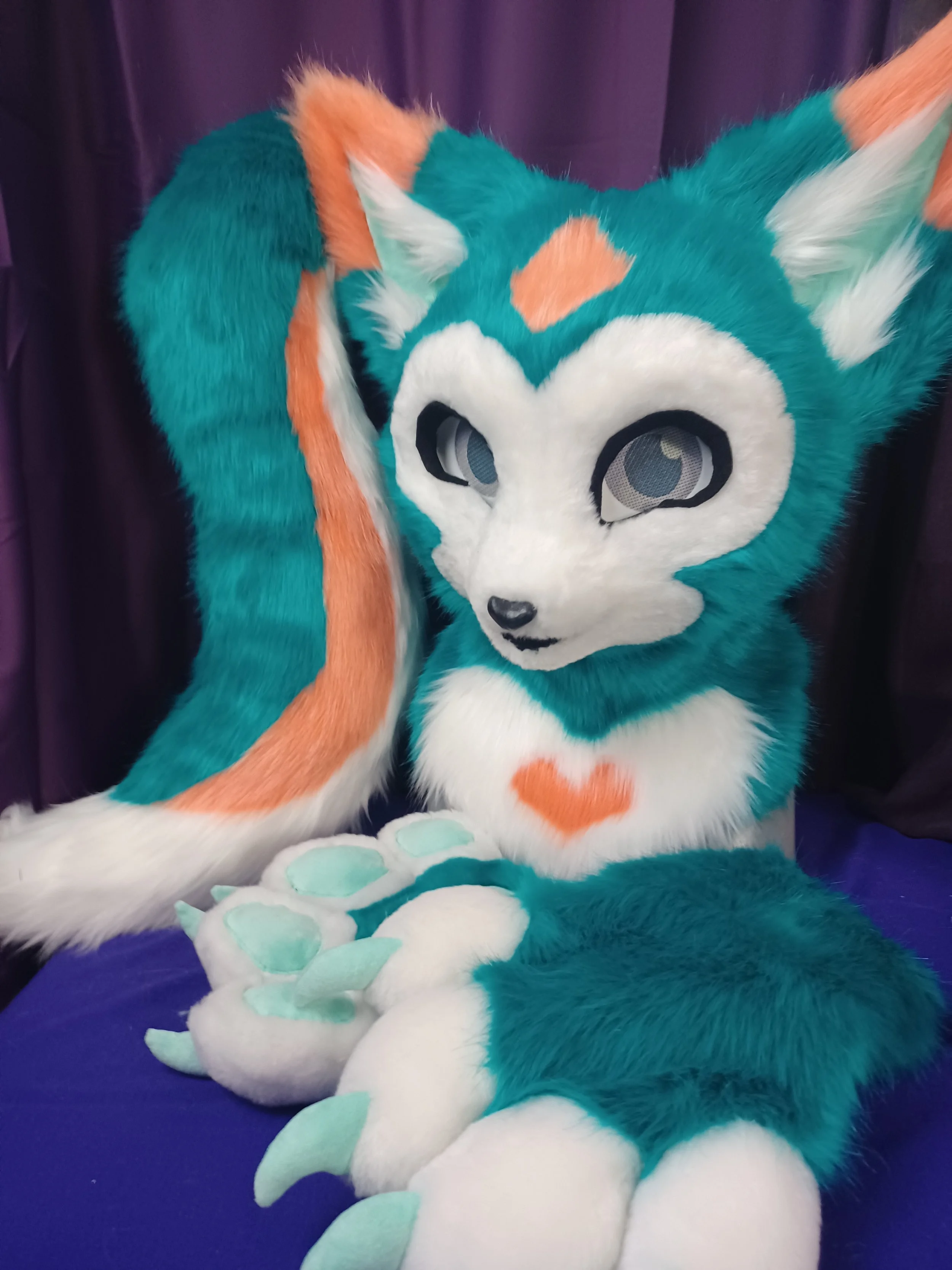 A person wearing a teal, orange, and white fursuit resembling a wolf or fox, with large eyes, sitting on a dark purple surface with a darker purple curtain background.