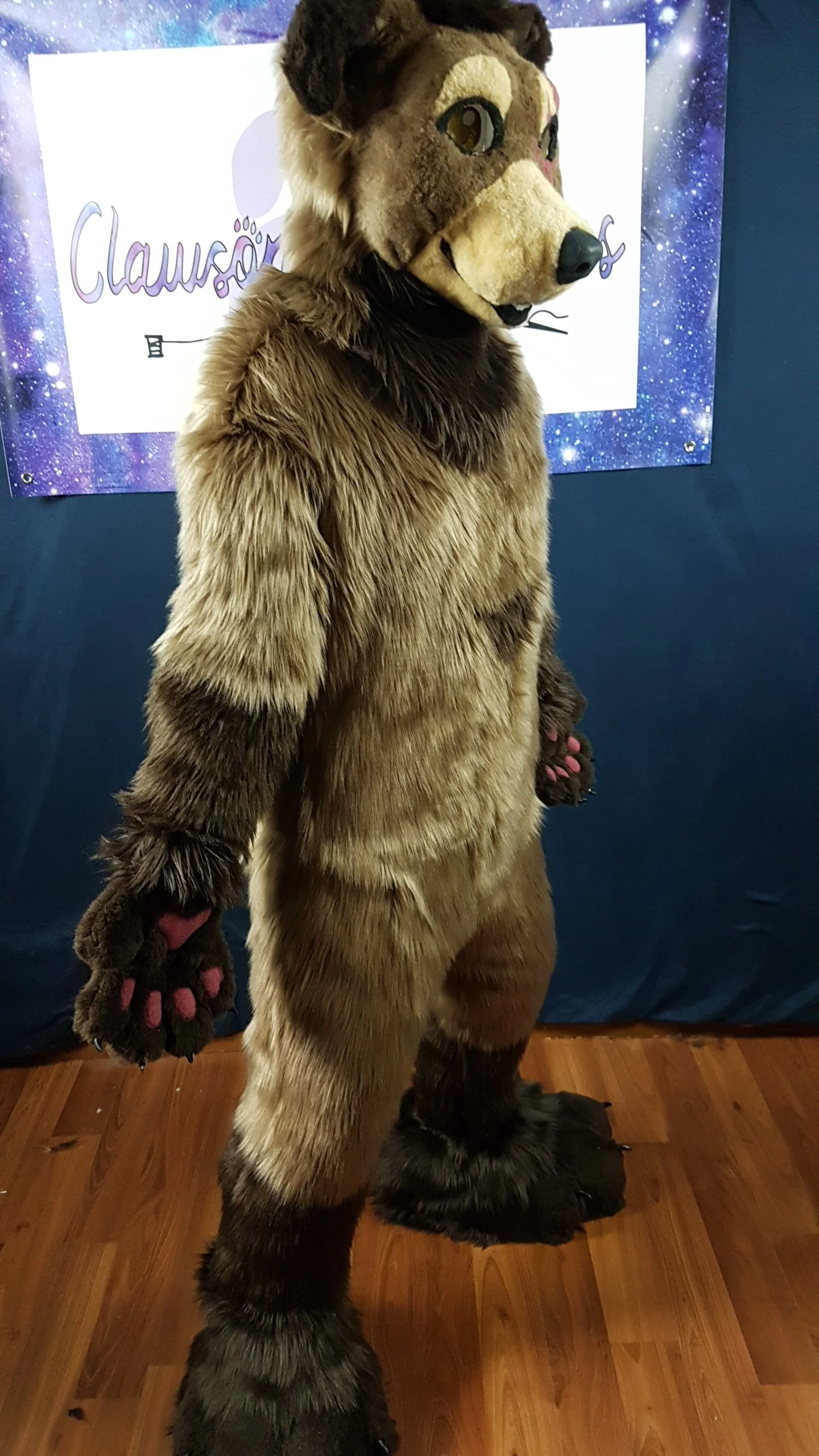 Person wearing a fursuit costume of a cartoonish wolf character, standing indoors with a backdrop of a space-themed banner.