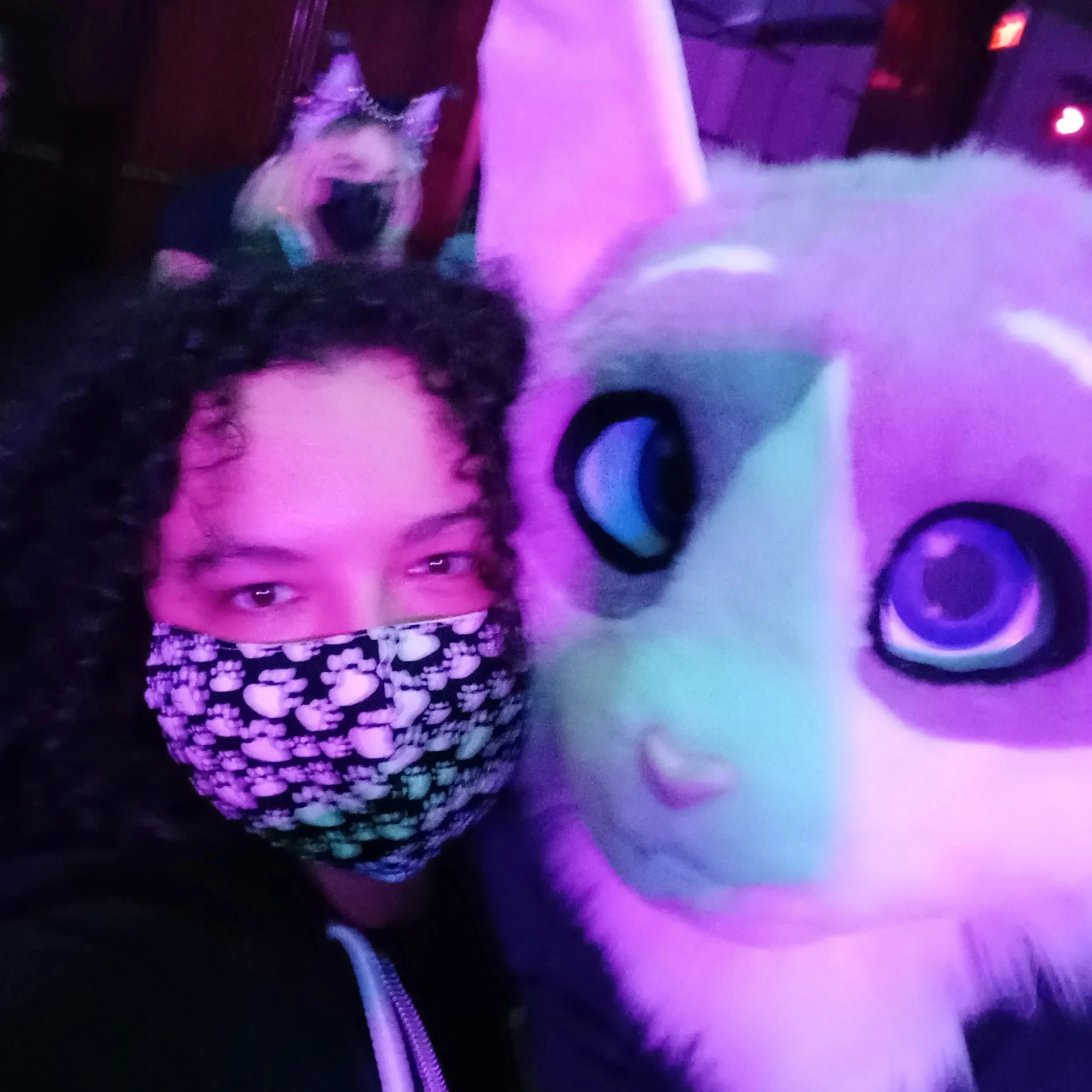 A person with curly hair wearing a patterned face mask stands in a dimly lit environment with purple and pink lighting.