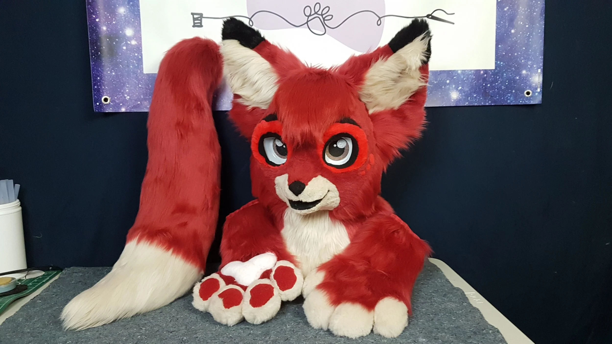 A person in a red fox mascot costume sitting on a table, with the mascot's head and full body visible. The background features a poster with a galaxy design and black line drawings. The mascot has large, expressive eyes, a white face with a black nos