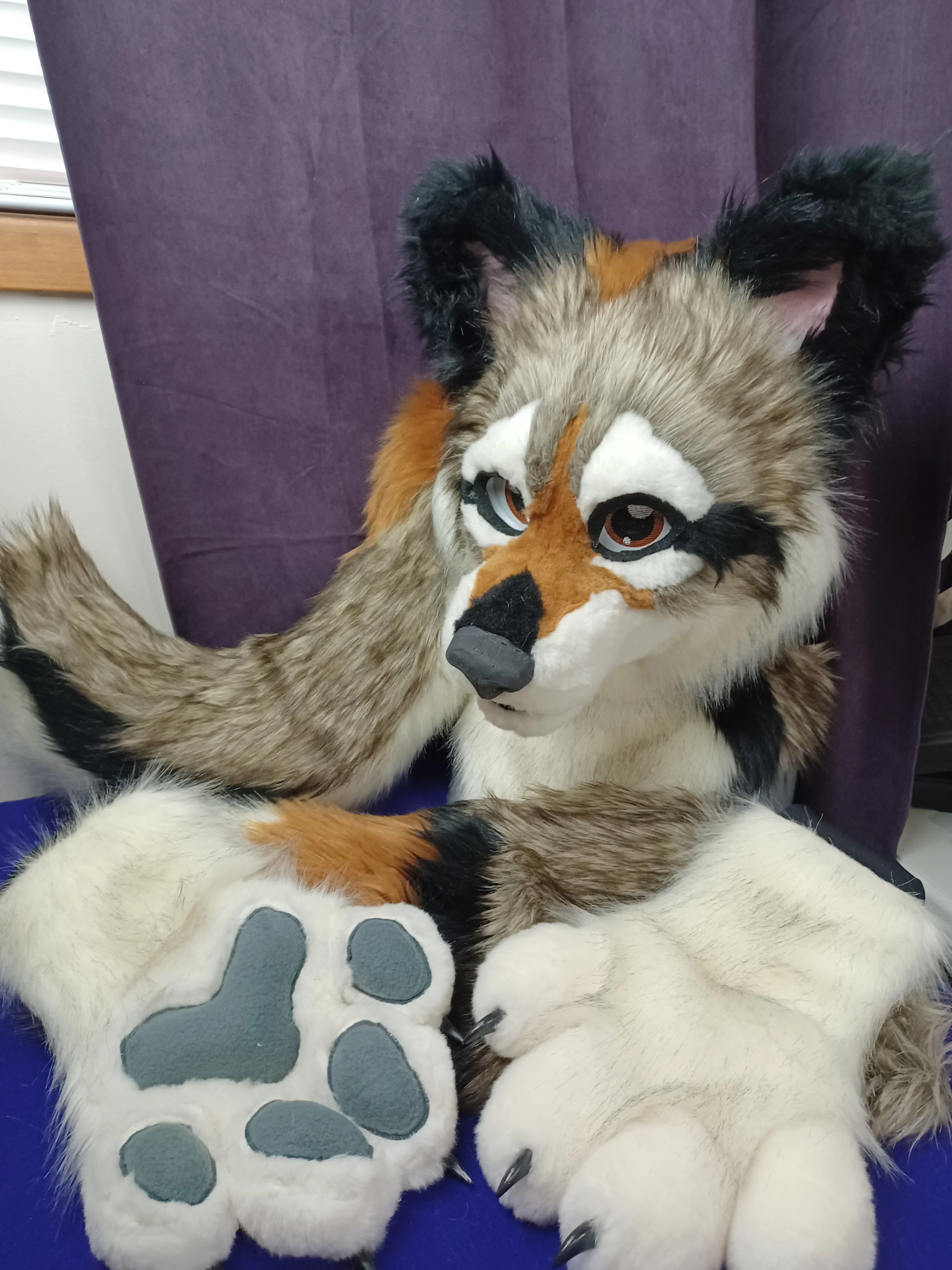 A person in a furry animal costume, resembling a fox or similar creature, sitting on a purple chair with their hand resting on the costume's paw. The costume features realistic fur and face details, including brown, white, black, and orange colors, w
