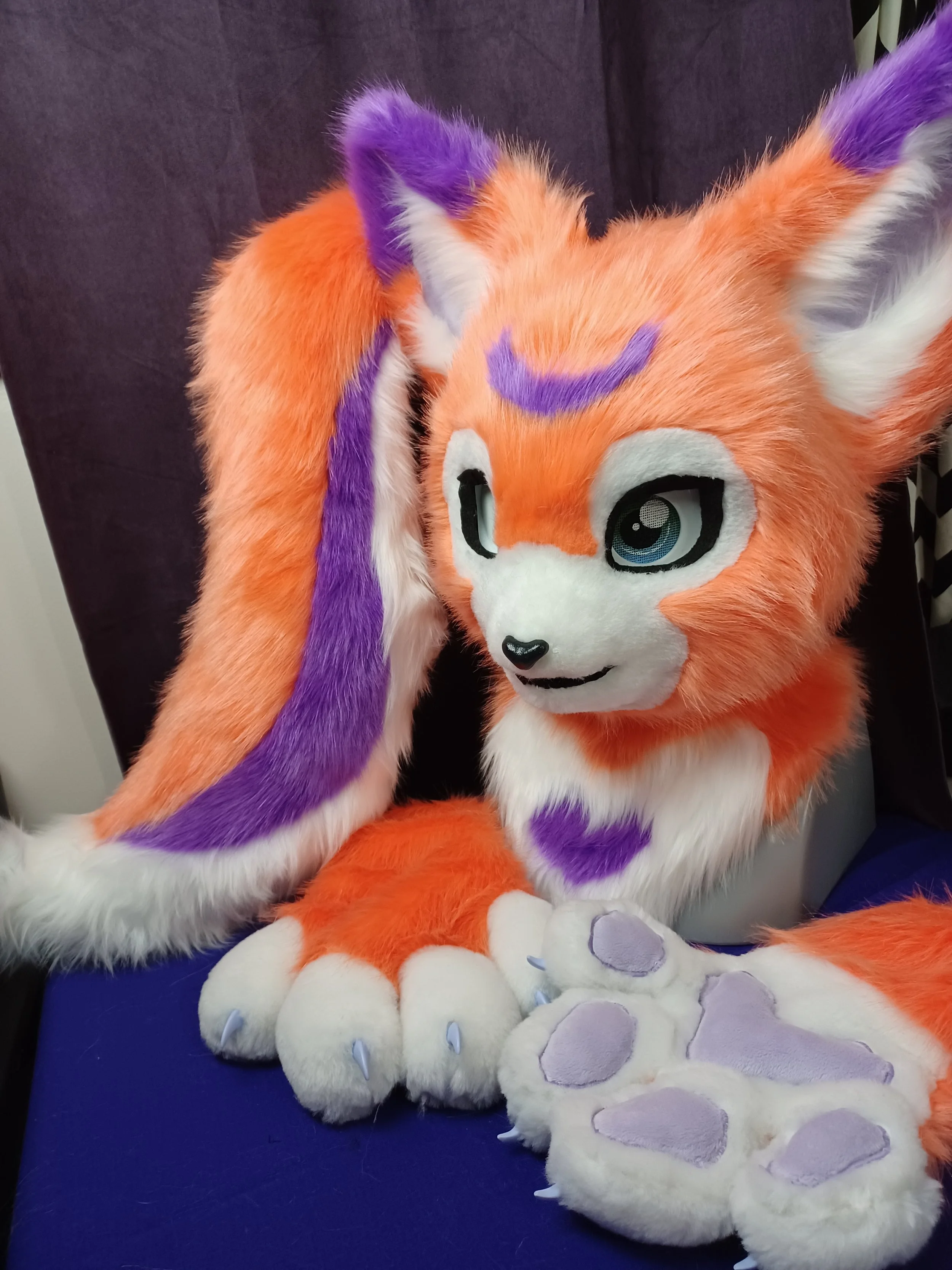A colorful furry animal costume resembling a fox, with orange, purple, and white fur, sitting on a blue surface with large plush paws in front of it.