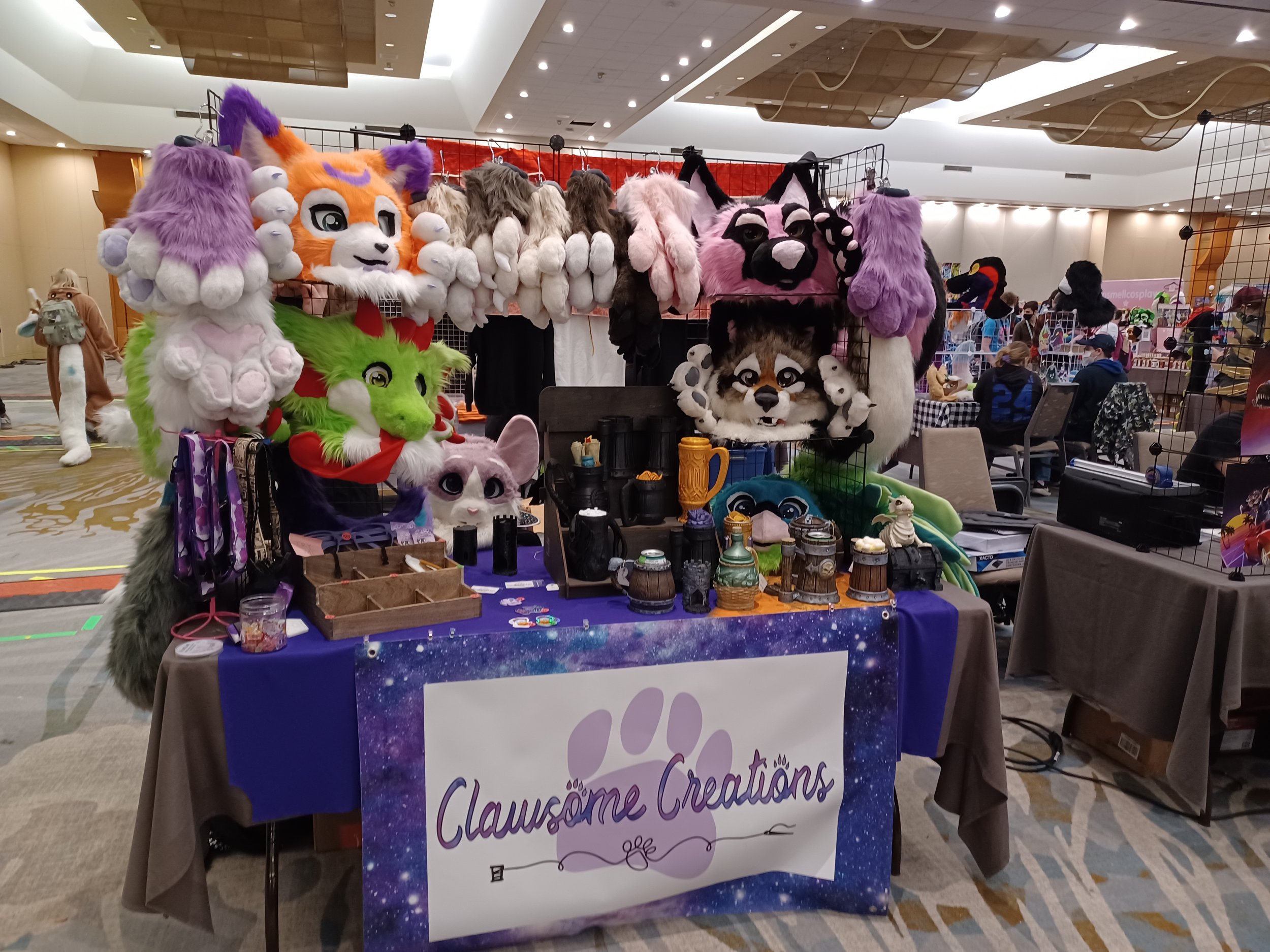 A booth with various plush animal masks and accessories displayed on wire racks at a convention with a sign with the words 'Clawsome Creations' is set up in a large indoor space with other booths and attendees visible in the background.