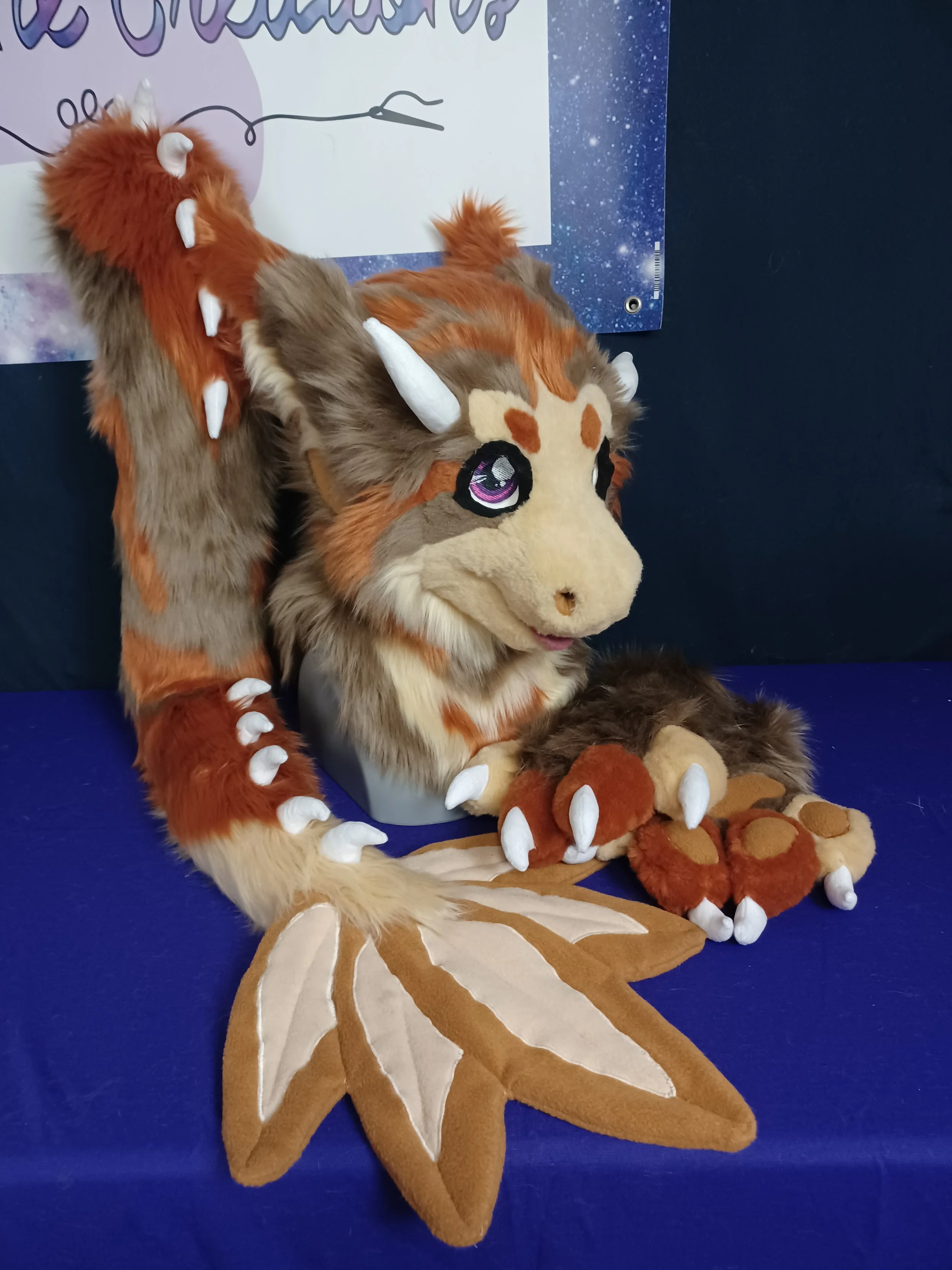 An angel dragon partial with a mane, horns, and large eyes, displayed on a purple cloth with a galaxy-themed poster in the background. the tail has a large splay of feathers at the tip