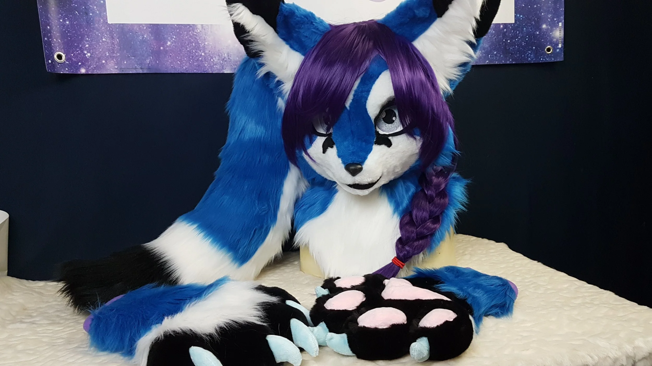 A colorful furry costume of a blue, white, and purple fox or wolf character with a long purple braid, sitting on a cream-colored surface against a dark background with a purple galaxy poster.