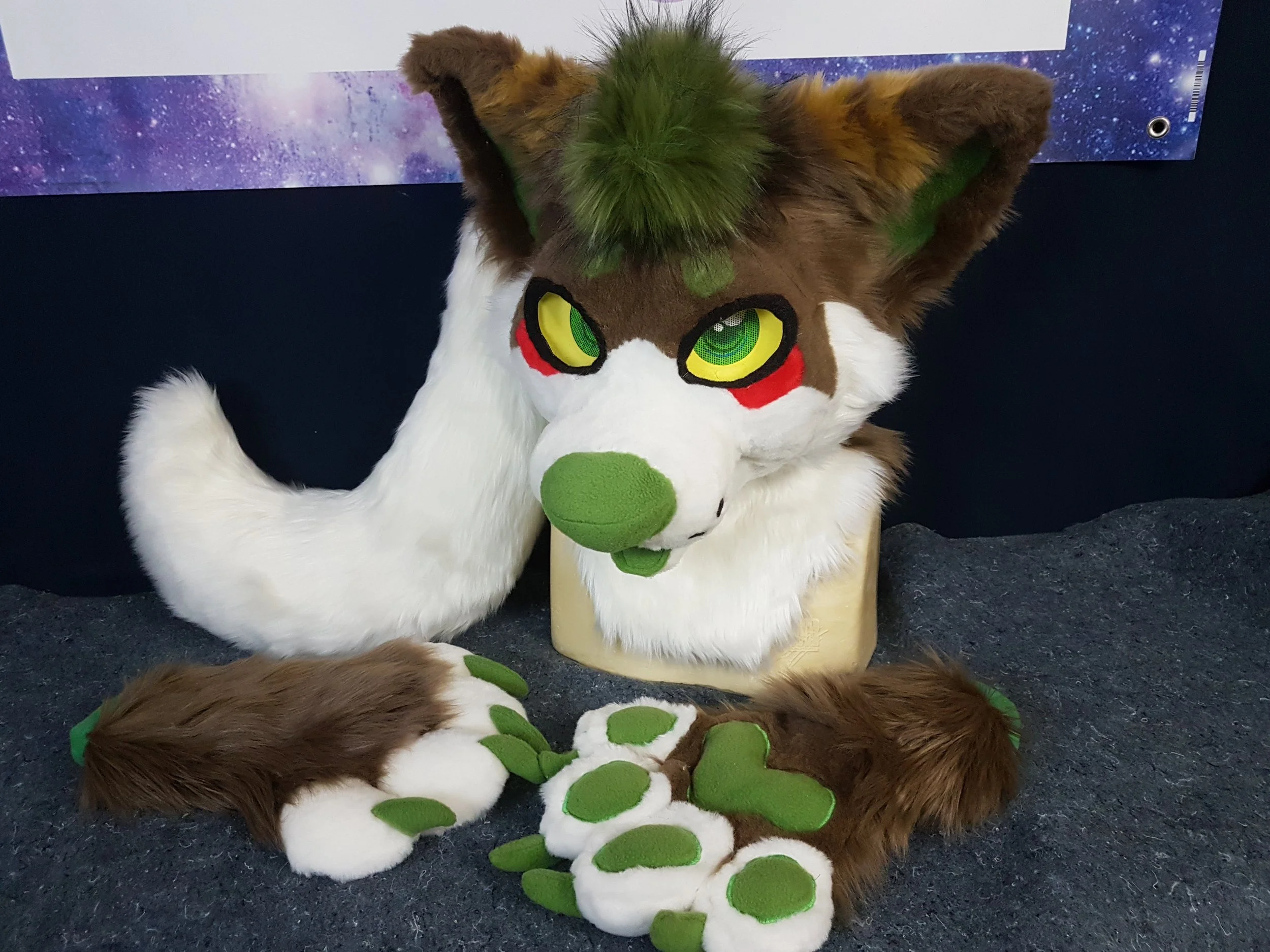A person in a furry animal costume with big yellow and green eyes, brown and white fur, and green spots on the face and paws, sitting on a dark surface near a space-themed poster.